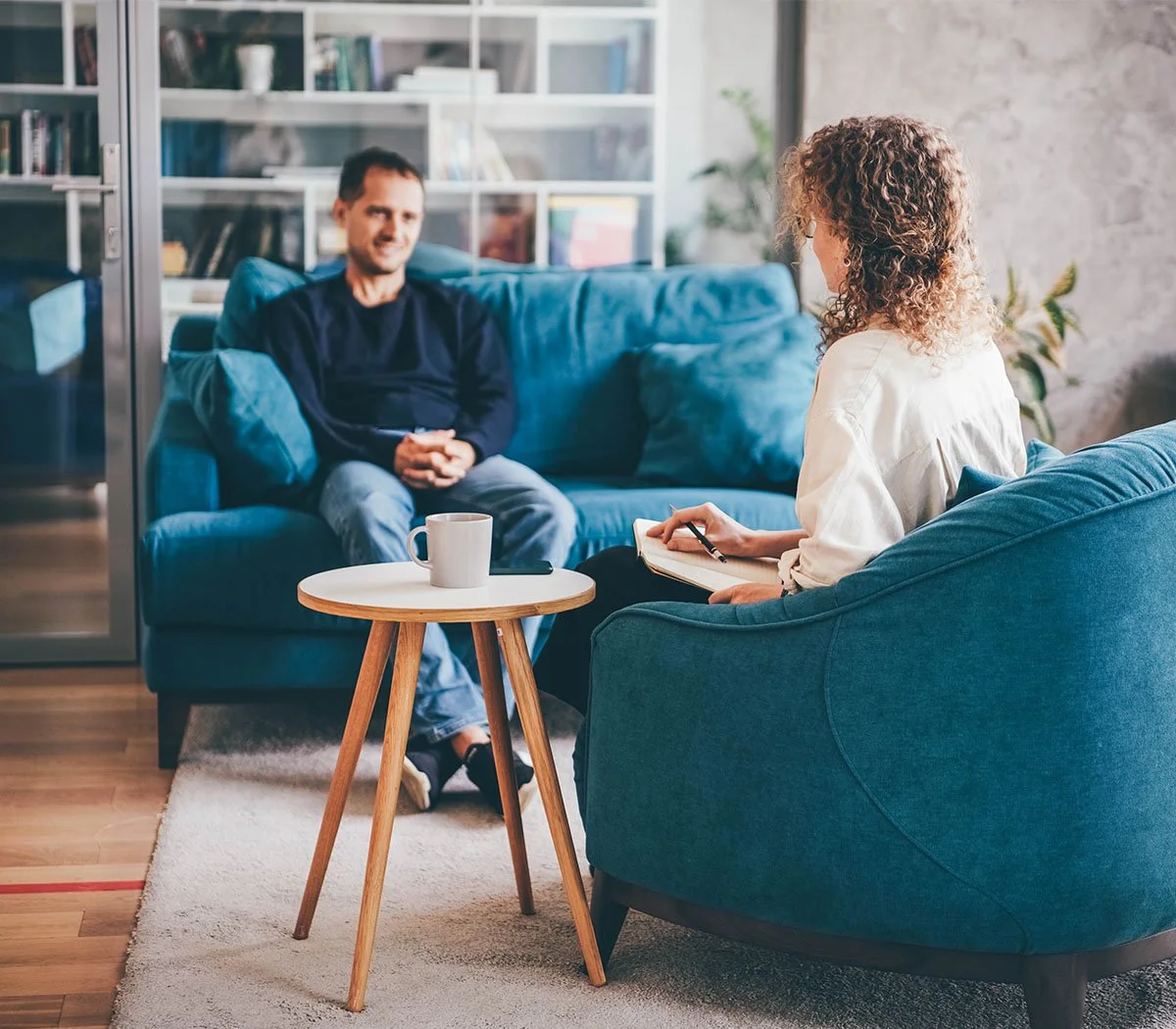 Zwei Personen in einem Gespräch in einem gemütlichen Raum, eine Frau sitzt auf einem blauen Sessel, hält einen Notizblock und schreibt, ein Mann sitzt auf einem blauen Sofa, beide wirken im Gespräch.