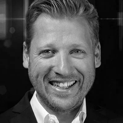 Schwarz-Weiß-Fotografie eines lächelnden Mannes in formeller Kleidung vor einem dunklen Hintergrund.