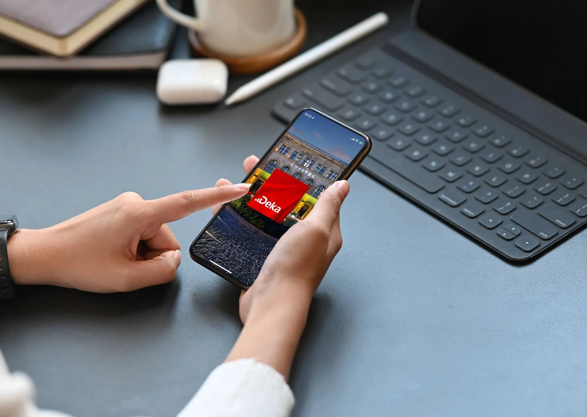 Eine Person hält ein Smartphone in der Hand, auf dem die Deka Bank Logo zu sehen ist, während sie an einem Schreibtisch mit Laptop, Notizbuch, Bleistift, AirPods und einem Becher sitzt.