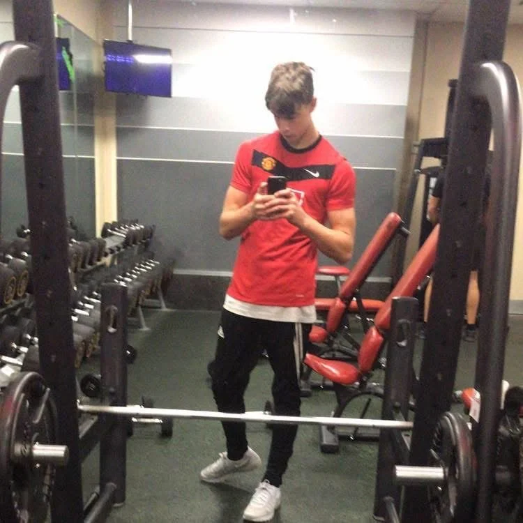 Young man in a red sports shirt, black pants, and white sneakers taking a mirror selfie in a gym, with workout equipment in the background.