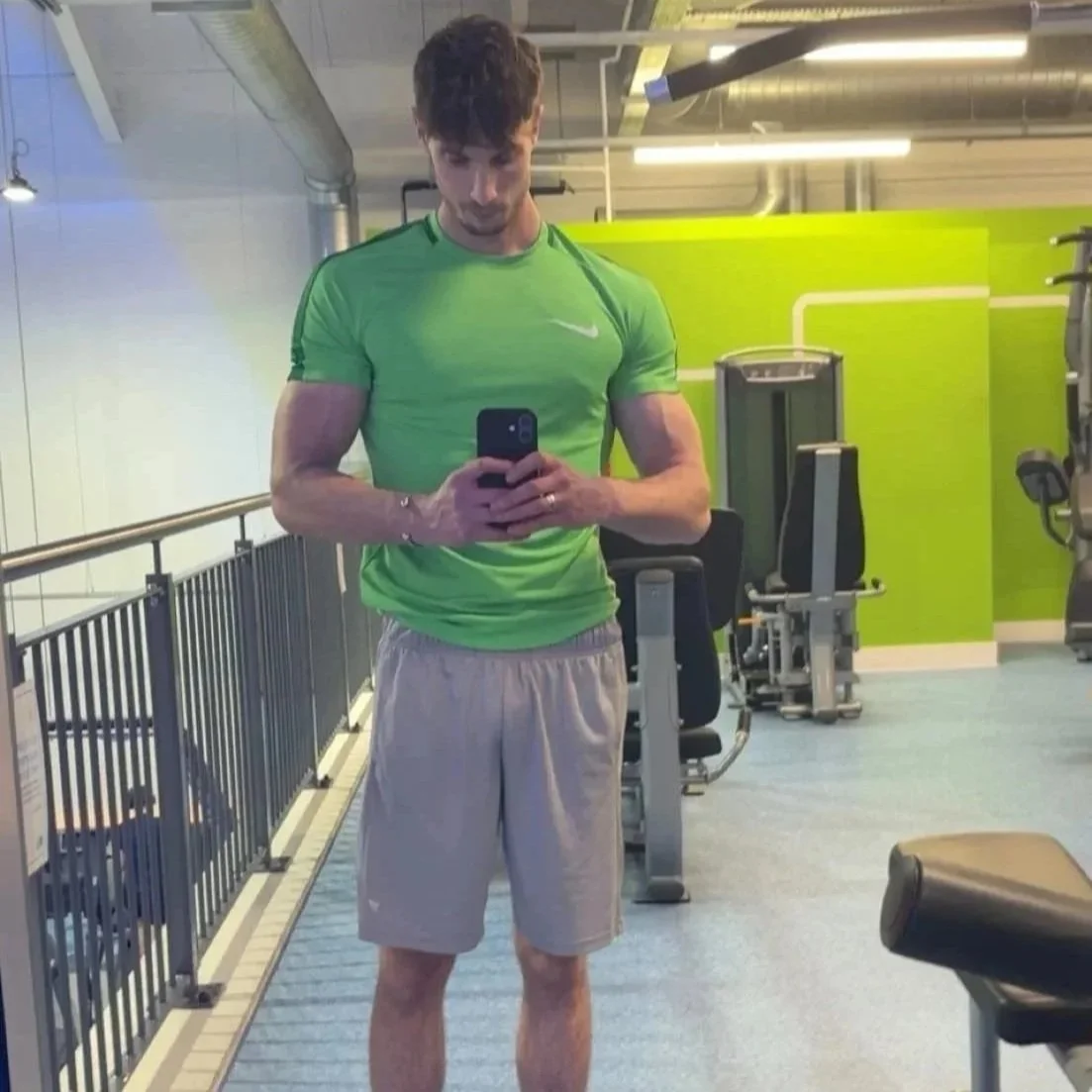 A young man in a green athletic shirt and beige shorts taking a selfie in a gym mirror.