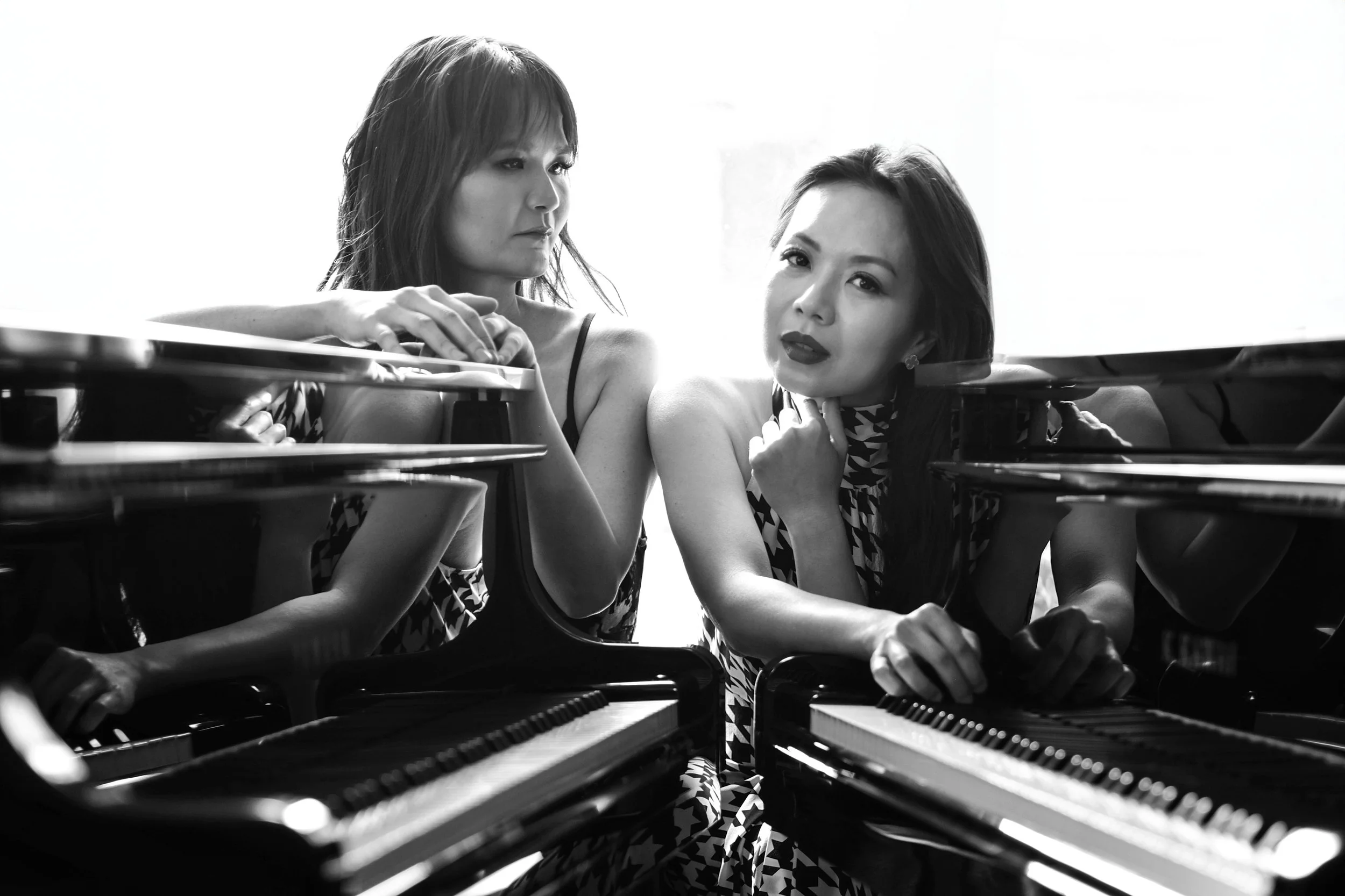 Two women in patterned dresses leaning on pianos, looking at each other in black and white photo.