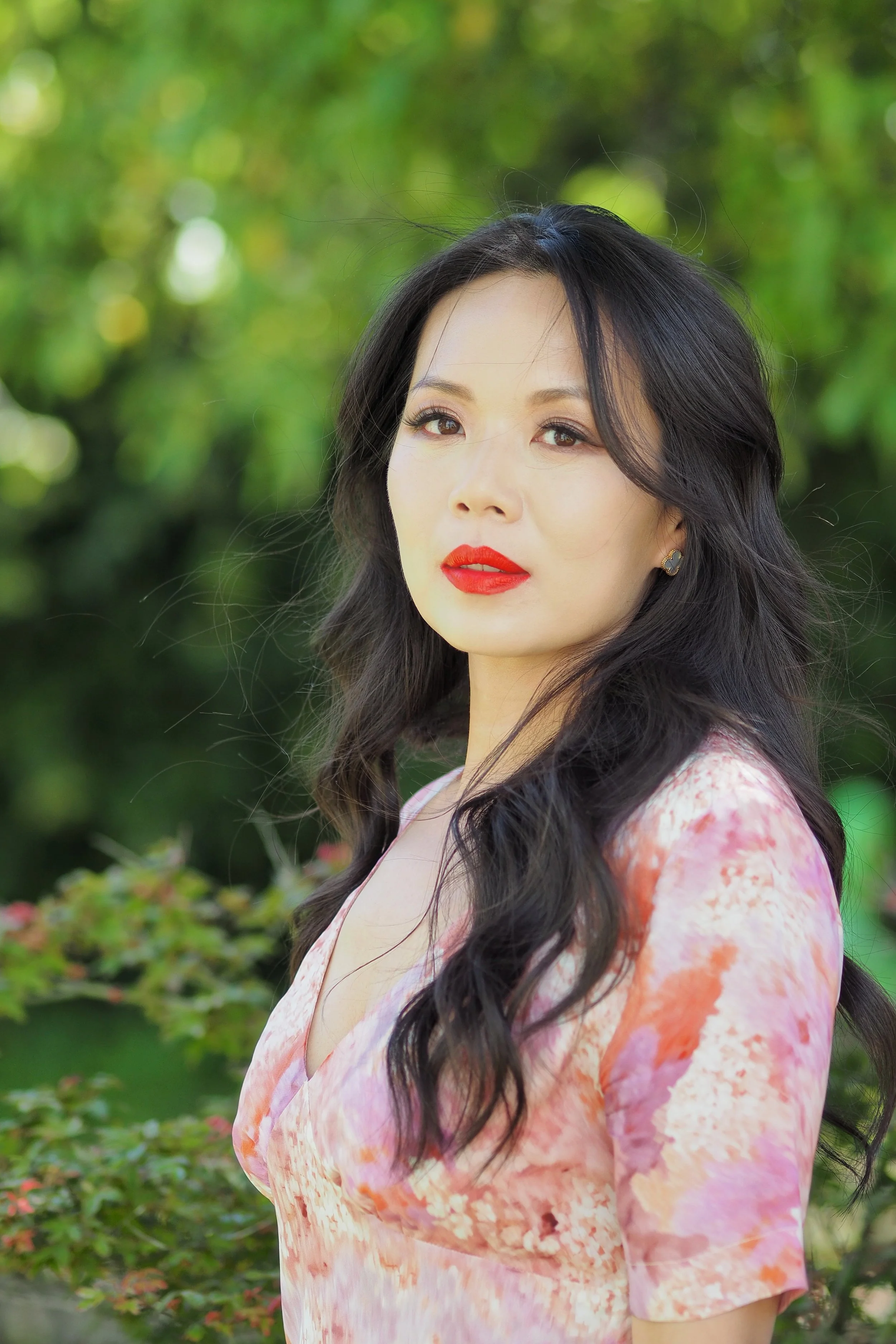 A woman with long, wavy black hair and red lipstick standing outdoors with greenery in the background.