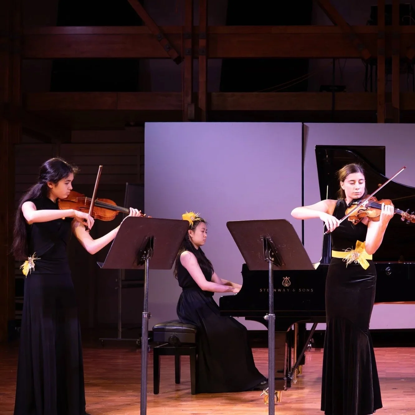 Three young women performing a classical music concert: two playing violins, one playing piano, all dressed in black with yellow accents, on stage with wooden floors and a dark background.