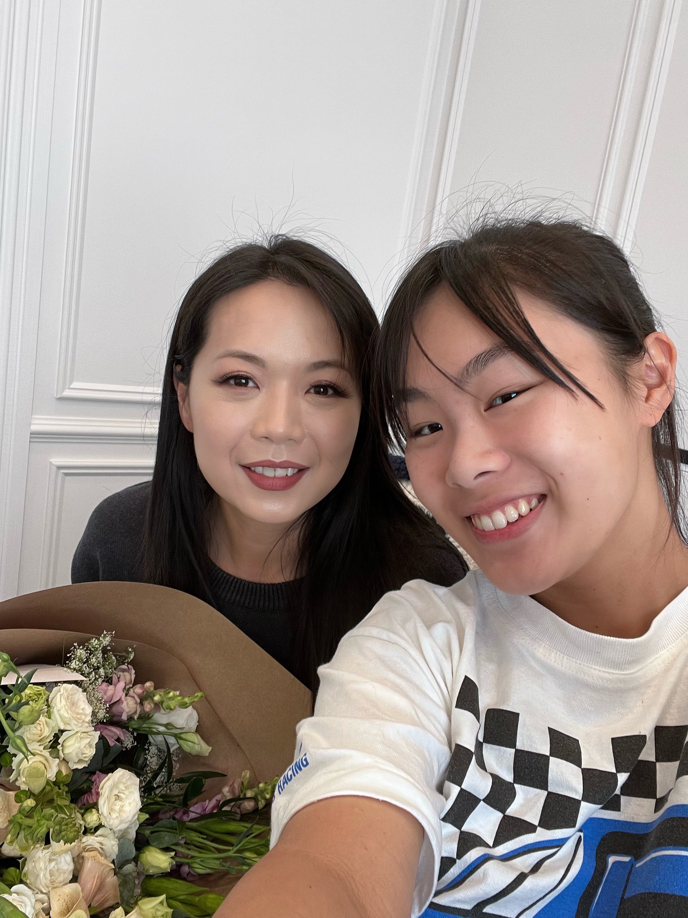 Two smiling women taking a selfie, with a bouquet of flowers in front of them, in a room with white paneled walls.