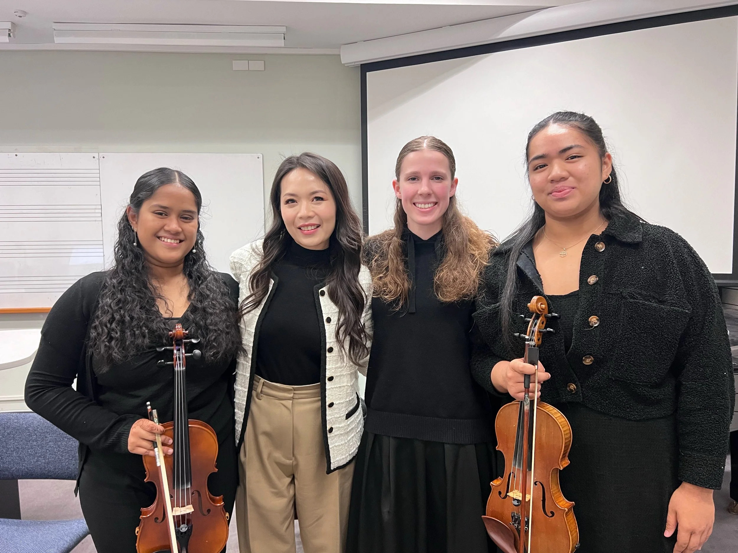 Four women standing together indoors, two holding violins, smiling at the camera.