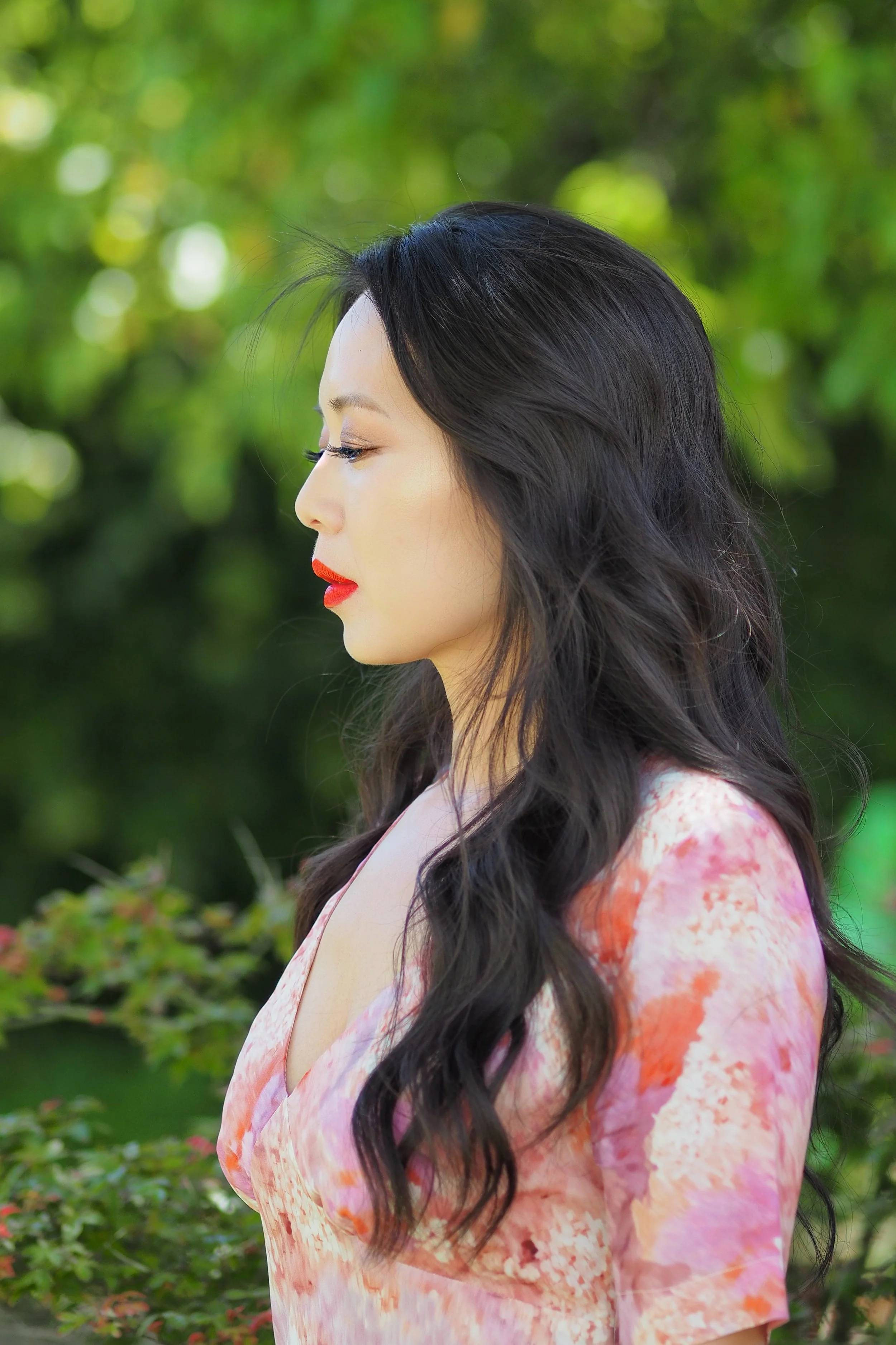 A woman with long, wavy black hair and red lipstick standing outdoors in front of green foliage, wearing a pink and orange floral dress.
