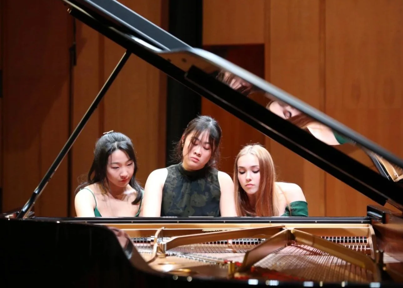 Three women sitting at a grand piano, with their eyes closed or looking down, appearing focused or emotional during a performance or rehearsal.