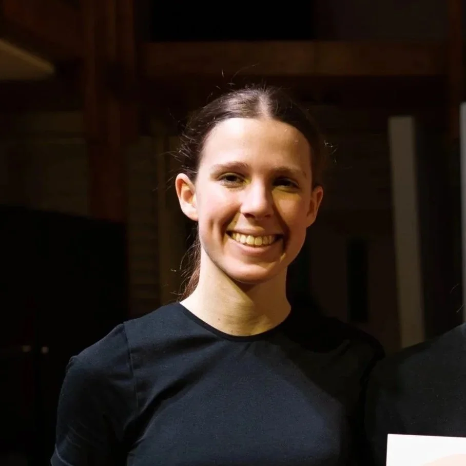 A young woman with brown hair tied back, smiling, wearing a black shirt, in a dimly lit indoor setting with wooden beams.