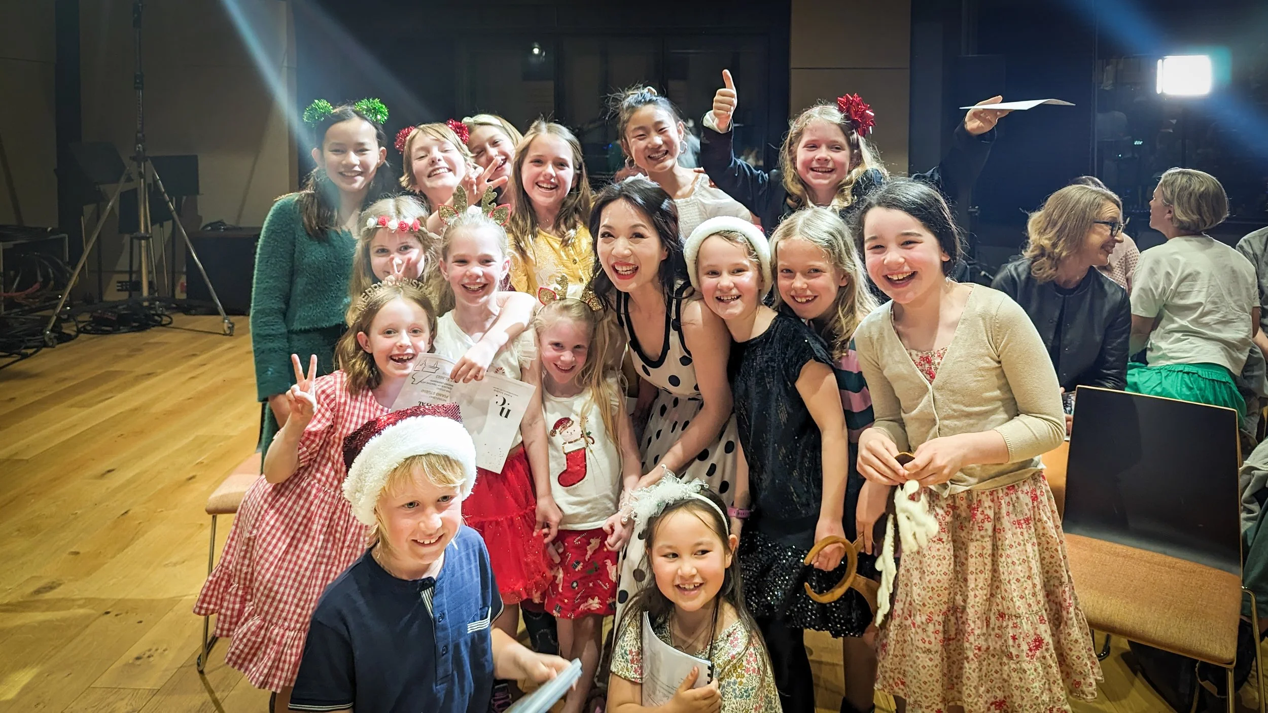 A group of smiling children and an adult woman posing together indoors, celebrating a holiday event, some wearing festive accessories like Santa hats and reindeer antlers.