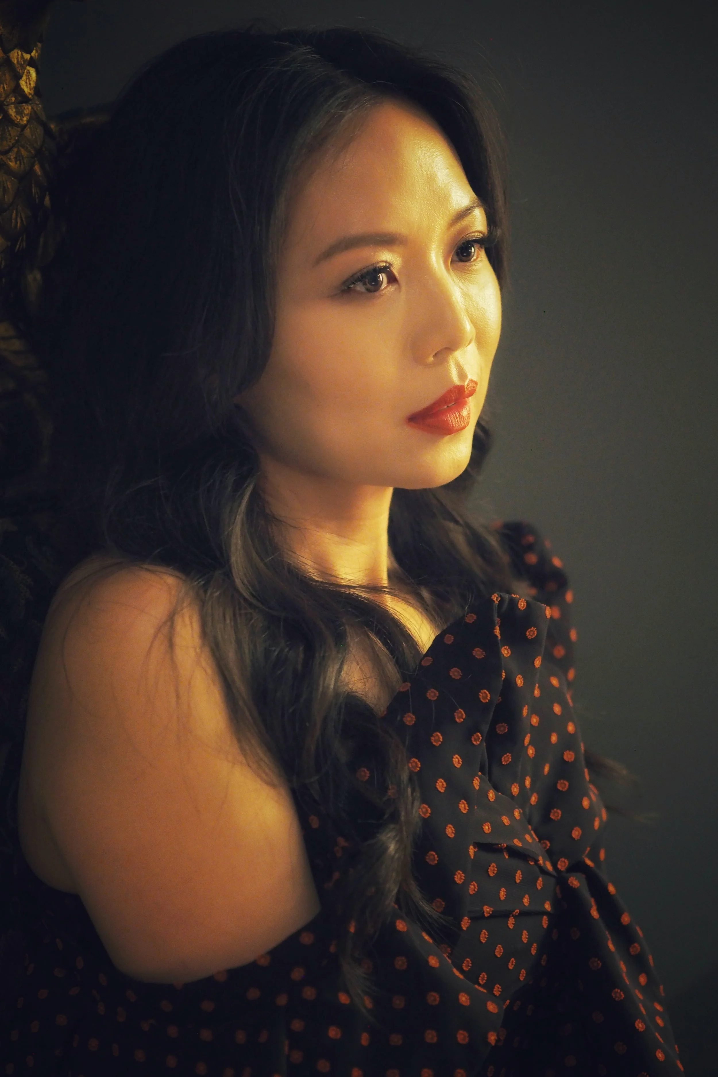 A woman with black hair and red lipstick wearing a black dress with orange polka dots, looking to the side against a dark background.