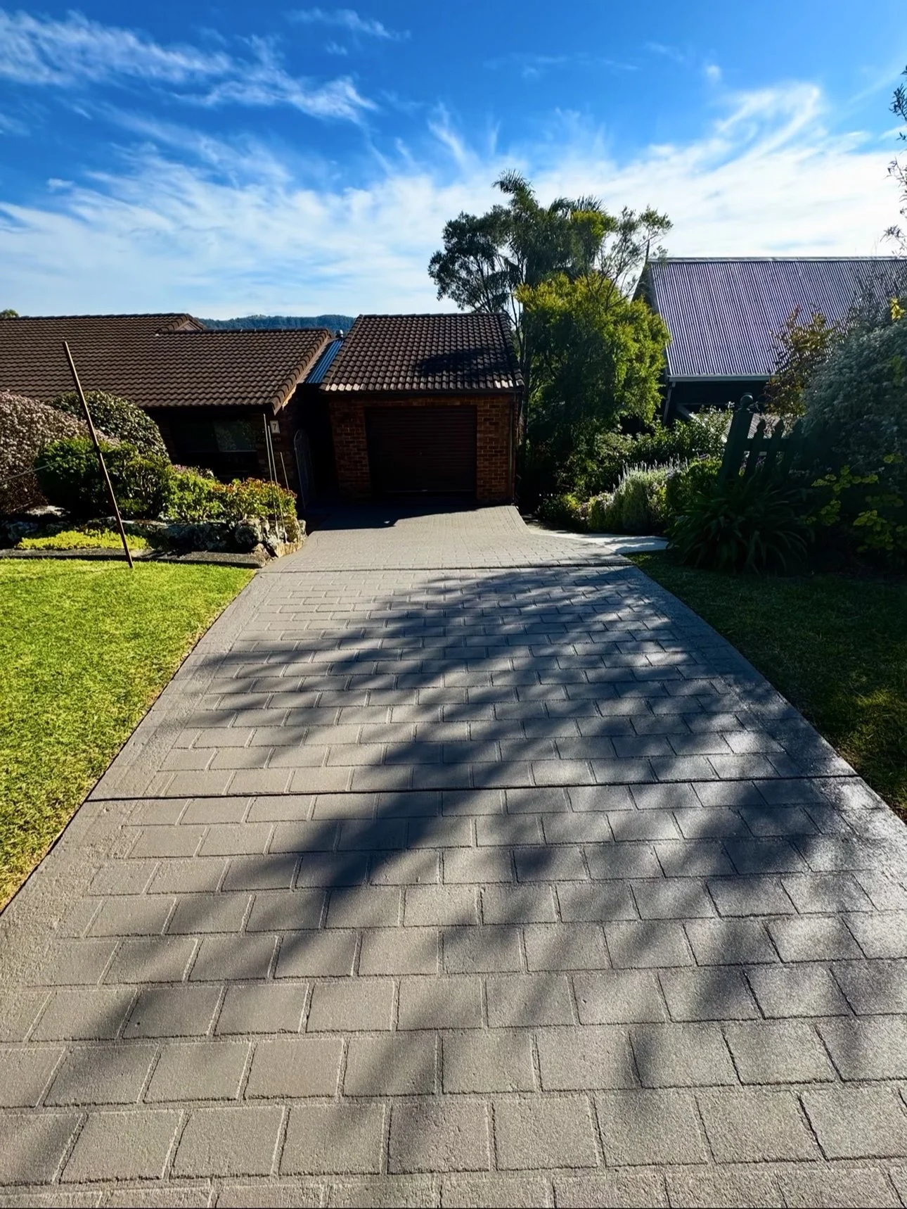 A paved driveway leading to a brick garage with a brown roof, surrounded by a green lawn and bushes. Trees and another building with a purple roof are visible in the background under a blue sky with some clouds.