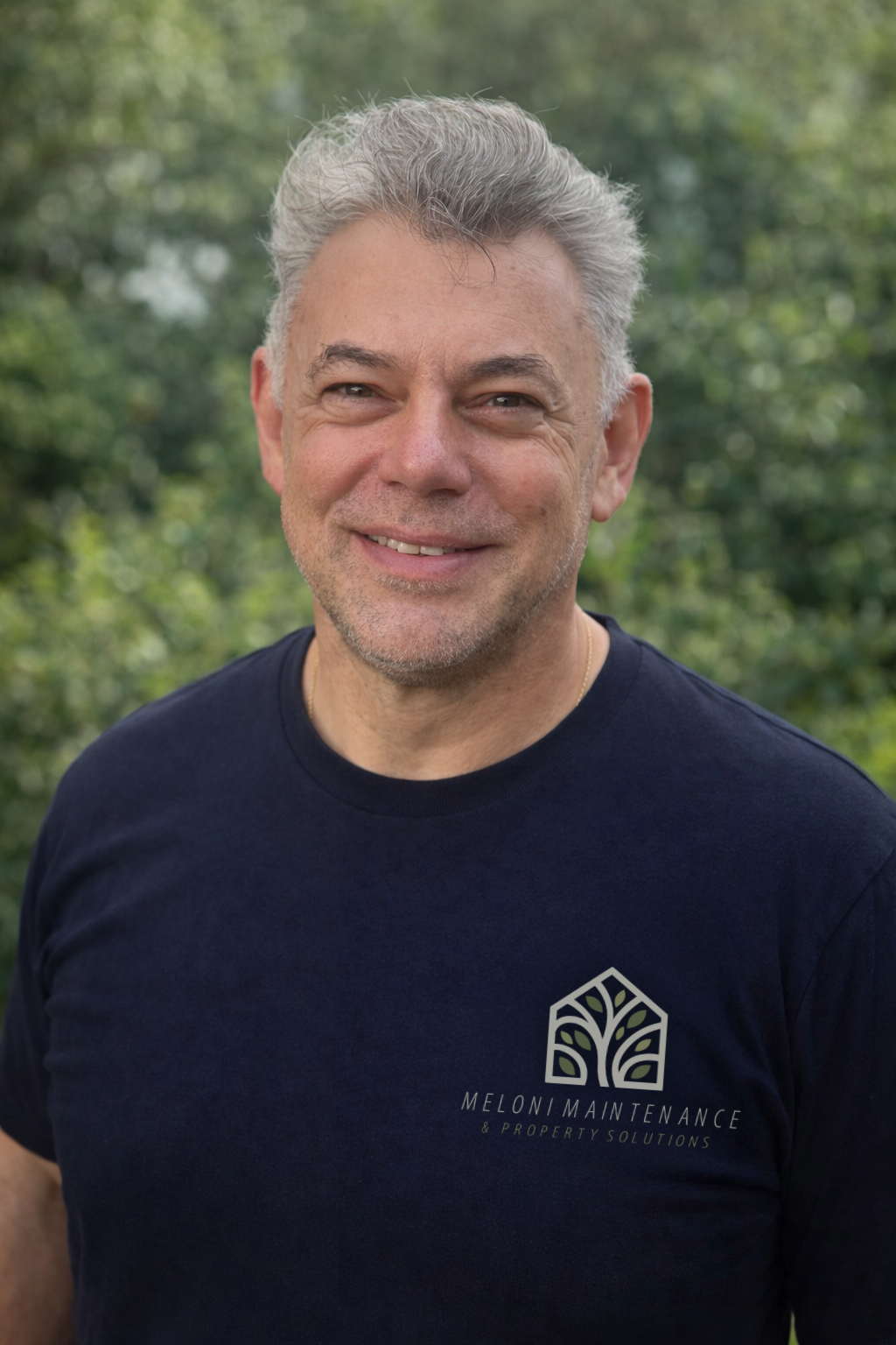 A smiling middle-aged man with gray hair, wearing a black t-shirt with a logo for Meloni Maintenance and Property Solutions, standing outdoors with green trees in the background.