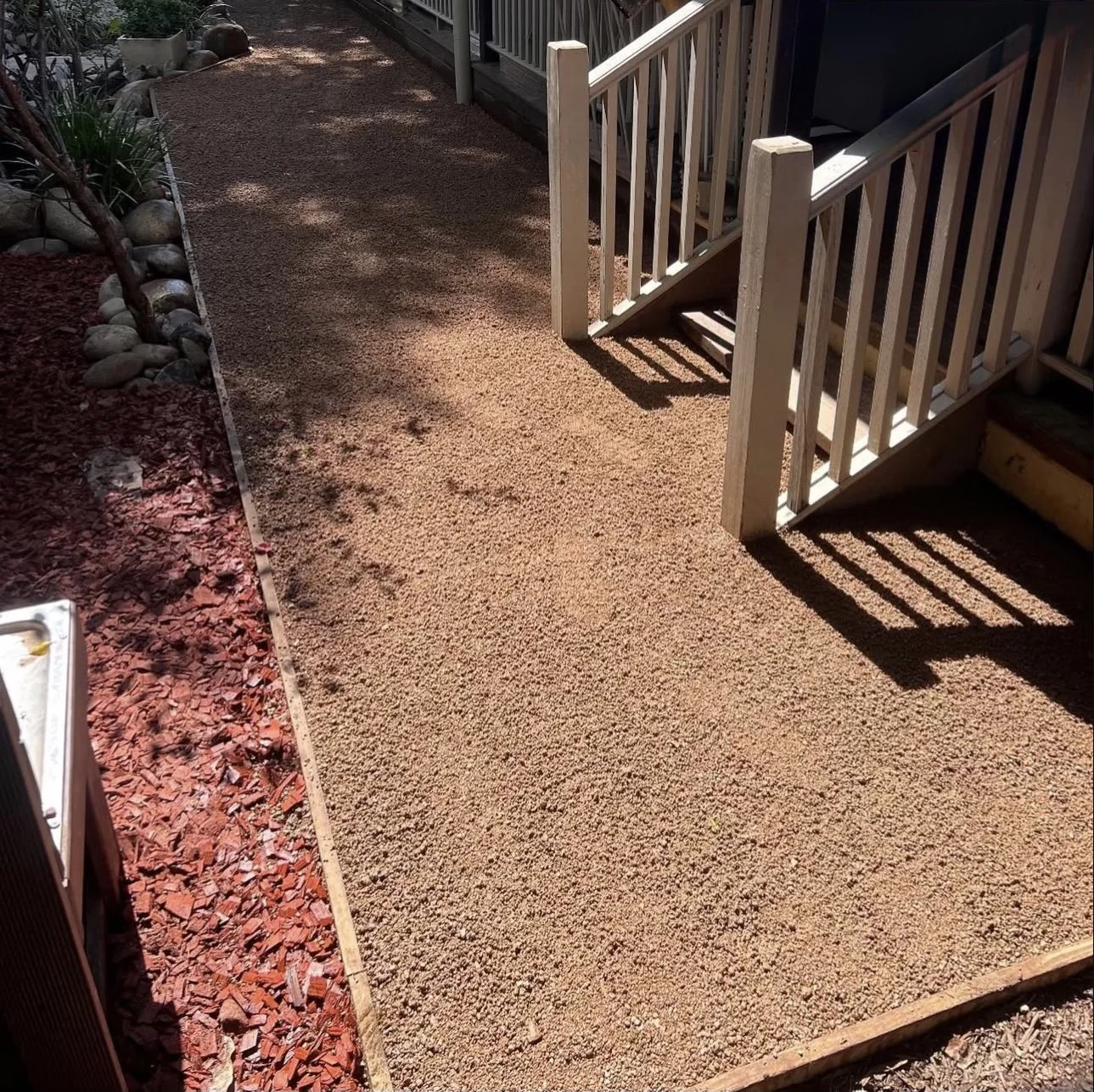 A backyard with a newly installed gravel pathway, bordered by red mulch and decorative rocks, with a wooden staircase leading to a deck.
