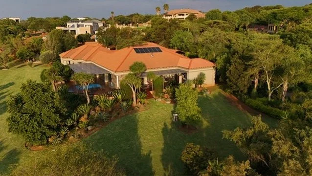 A large house with a red-tiled roof and solar panels, surrounded by lush green trees and a spacious lawn with various plants and bushes.