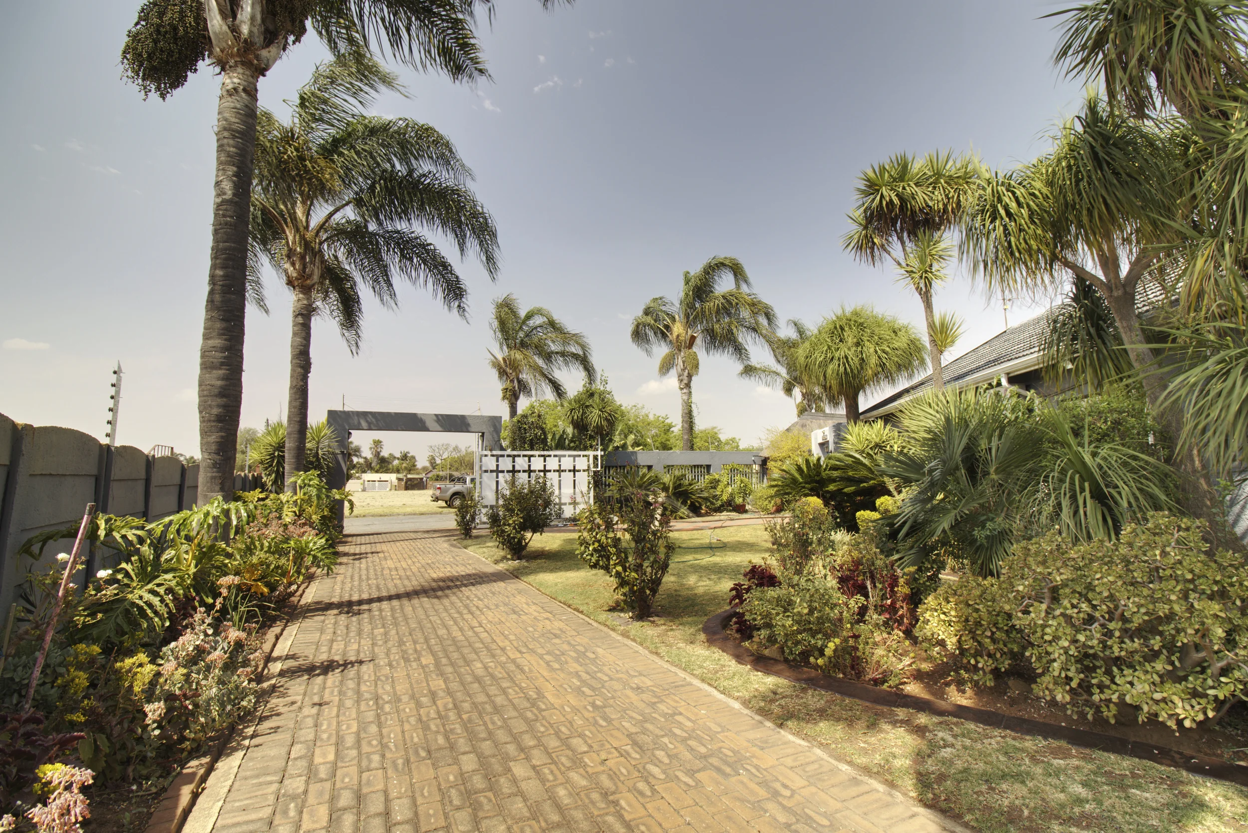 A landscaped garden with a paved walkway, various lush plants and trees, including palm trees, and a building with a tiled roof in the background, under a mostly clear sky.