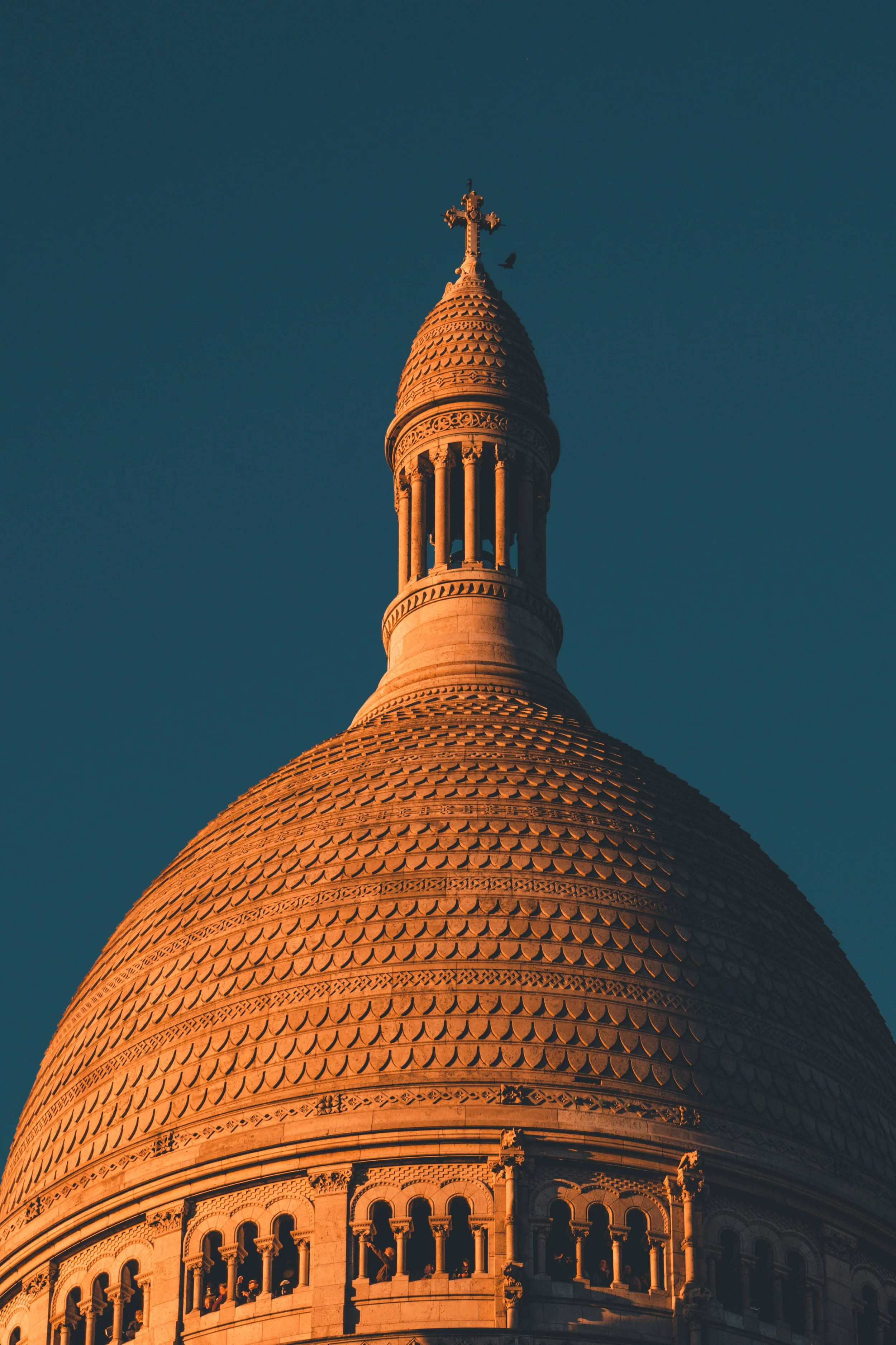 Une photographie de la tour de La Basilique du Sacré-Cœur de Montmartre, Paris, capturée lors du coucher du soleil avec la lumière dorée. La tour est ornée de détails architecturaux et un oiseau vole près du sommet.