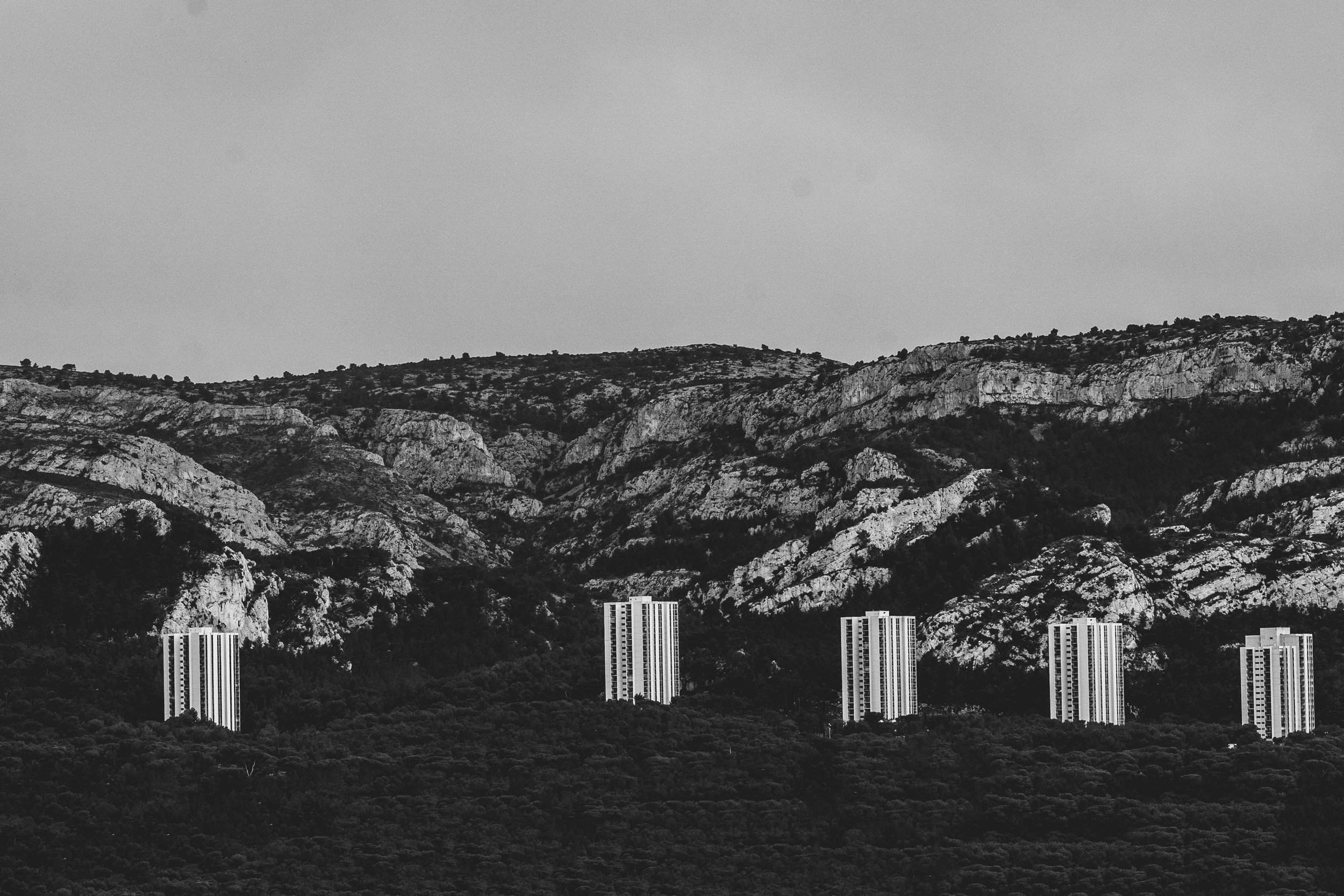 Cinq immeubles hauts devant une montagne rocheuse avec une végétation dense, en noir et blanc.