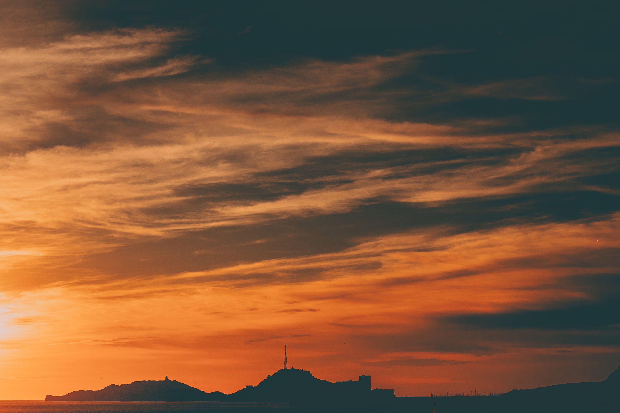 Coucher de soleil sur la mer avec des clouds colorés dans le ciel, silhouette d'une île ou d'une péninsule avec une tour ou antenne sur une colline.