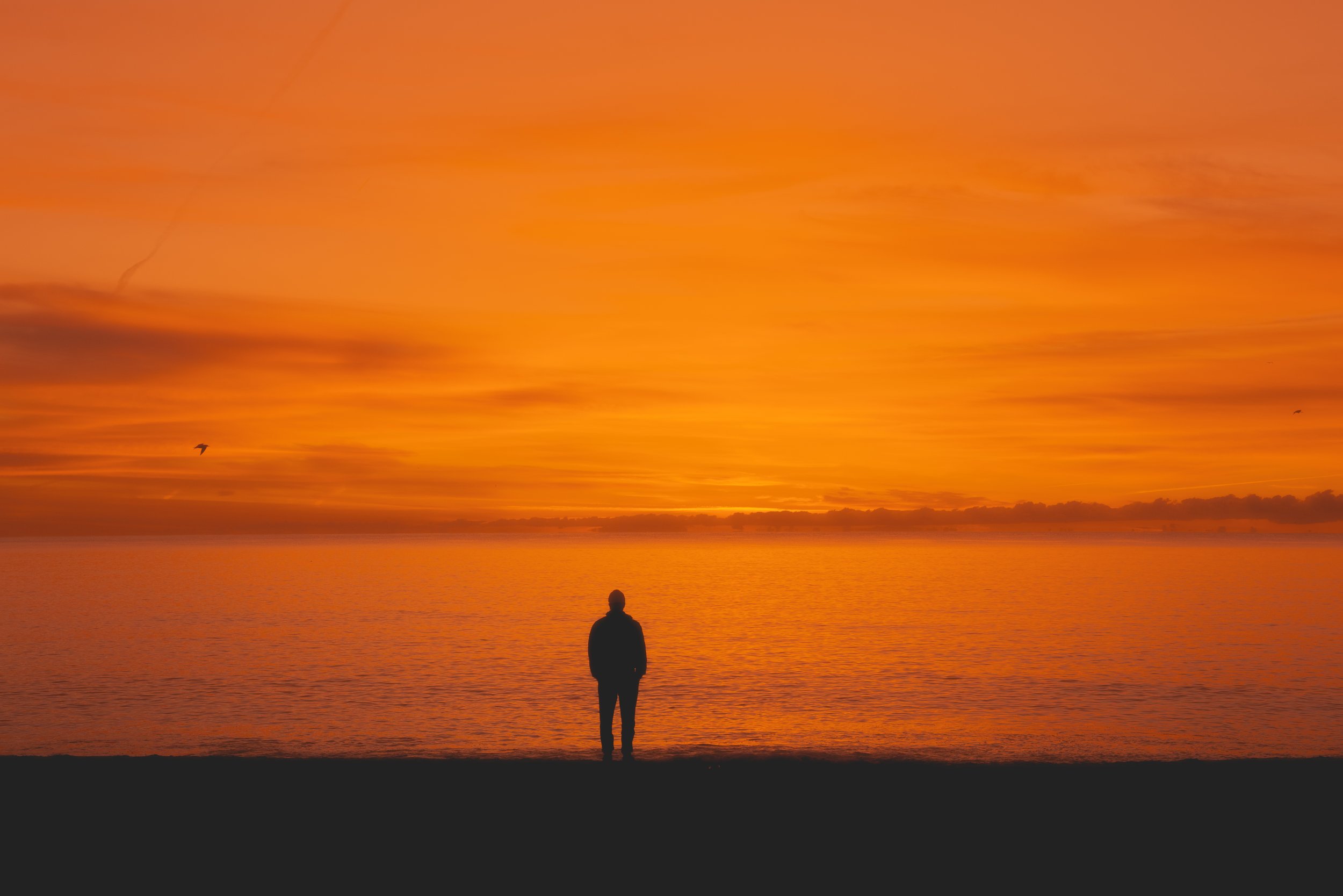 Silhouette d'une personne debout sur la plage regardant un coucher de soleil orange sur la mer.