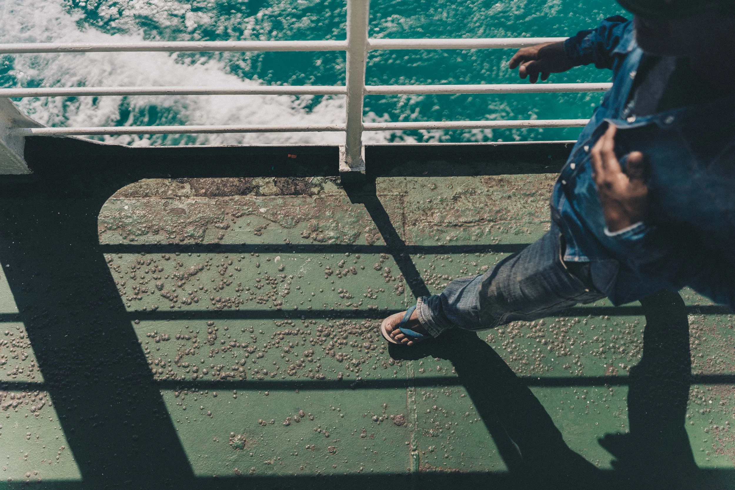 Une personne marche sur le pont d'un bateau ou d'un ferry, vue du dessus, avec un sol vert et des rails blancs, en regardant les eaux en dessous.