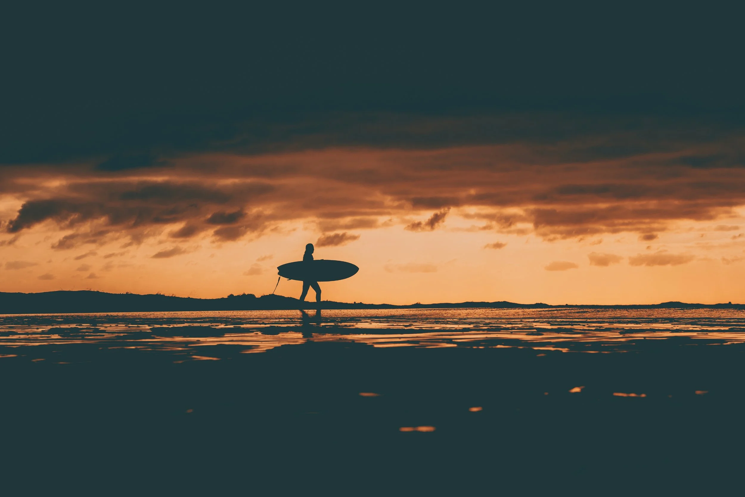Surfeur marchant sur la plage au coucher du soleil avec un surf sous le bras, silhouette contre un ciel orageux.