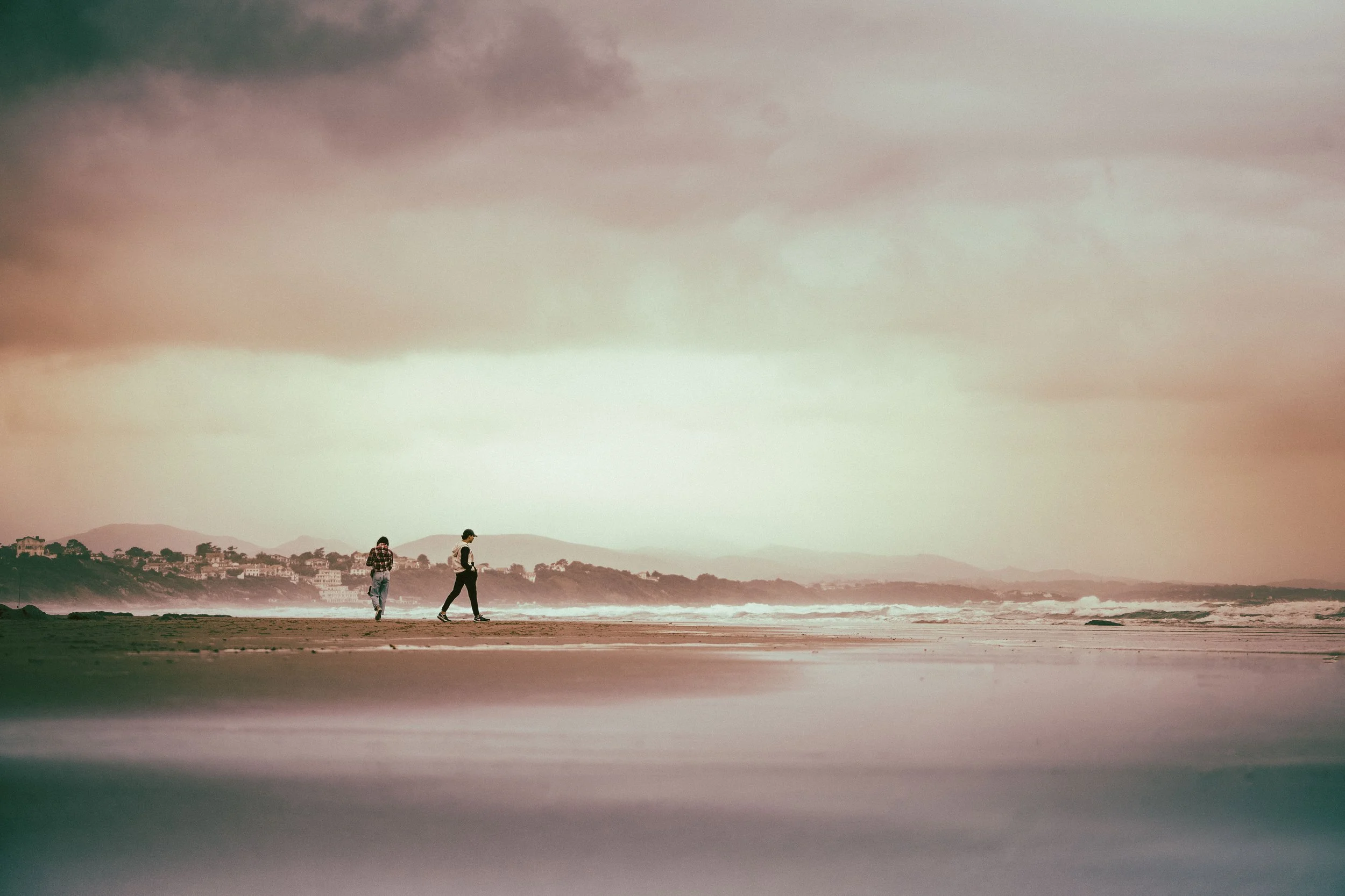 Deux personnes marchent sur une plage près de l'océan, avec des montagnes au loin, sous un ciel nuageux.