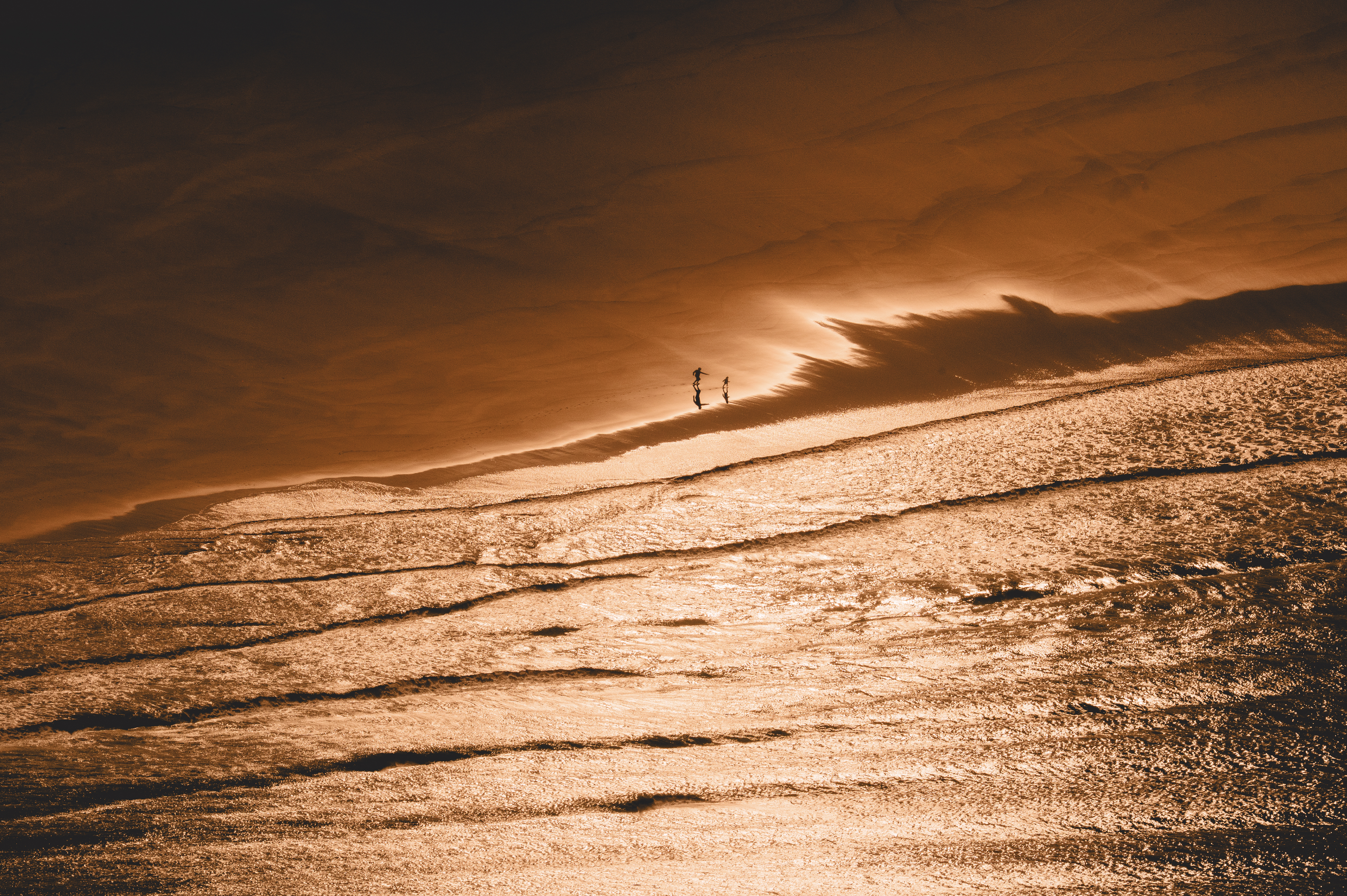 Deux personnes marchant sur la plage au coucher du soleil, avec des vagues éclairées par la lumière orangée.