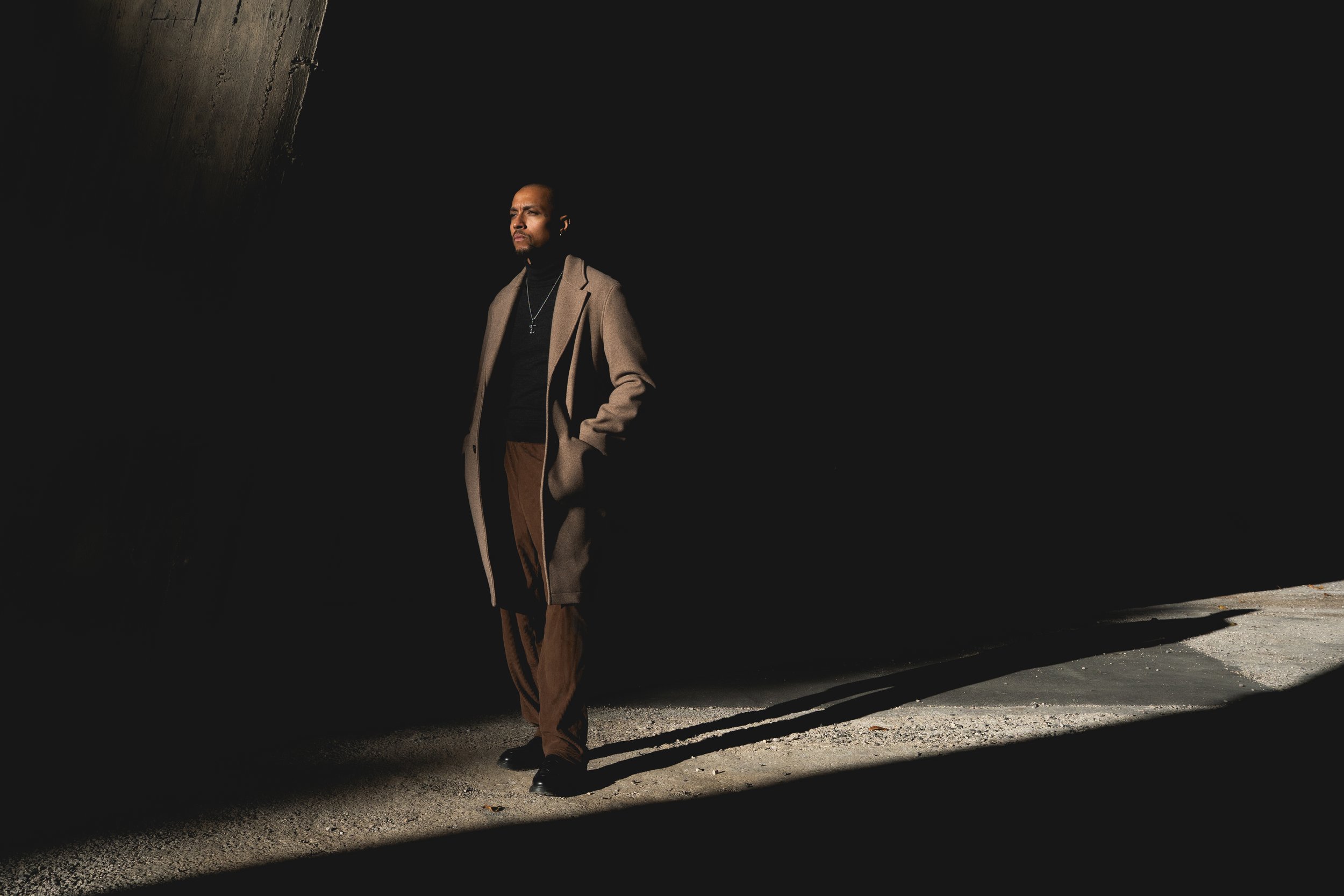 Homme en manteau beige, t-shirt noir et pantalon marron, debout dans une zone sombre avec un rayon de lumière sur lui, avec un fond obscur et une ombre allongée au sol.
