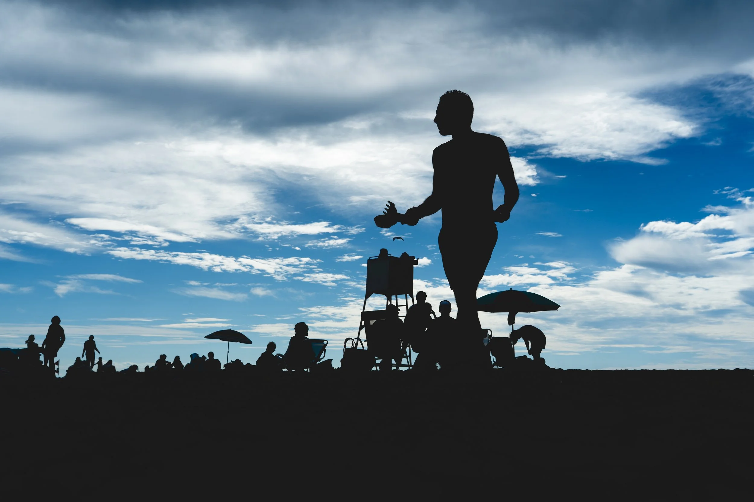 Silhouettes de personnes sur une plage au coucher du soleil, certaines sous des parasols, avec un ciel orageux et nuageux en arrière-plan.
