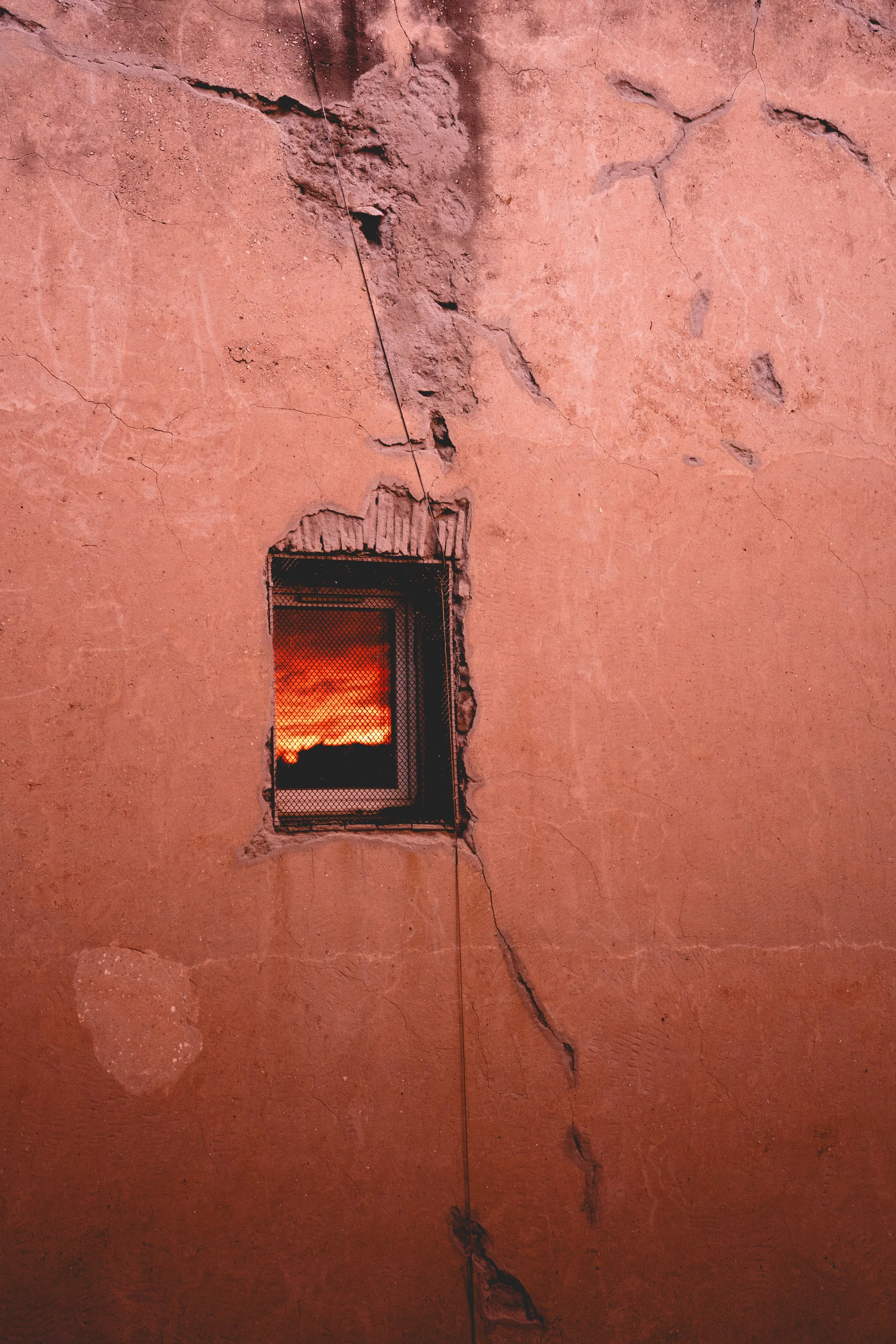 Une vieille façade en pierre ou en plâtre avec une fissure importante. Il y a une petite fenêtre avec une grille, laissant voir un coucher de soleil avec un ciel orange et rouge. La façade est de couleur rose ou ocre.
