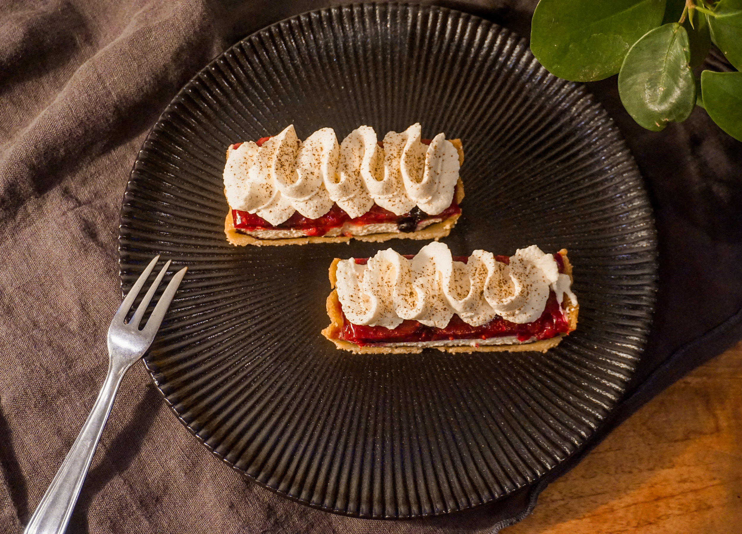 Zwei Frucht-Tartes mit Streuseln und Schlagsahne auf einem schwarzen Teller, daneben ein Messer und ein Zweig mit grünen Blättern auf einem braunen Stoffuntergrund.