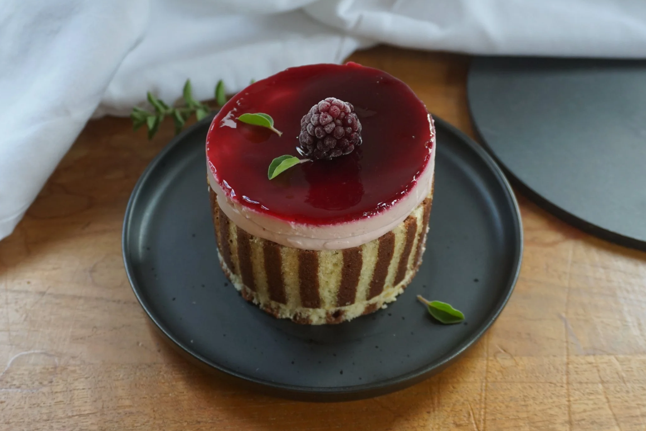 Kuchen mit rotem Fruchttopping, Brombeere und grünen Blättern auf einem schwarzen Teller, auf einem Holztisch. Im Hintergrund weiße Stoffserviette und dunkler Stein.