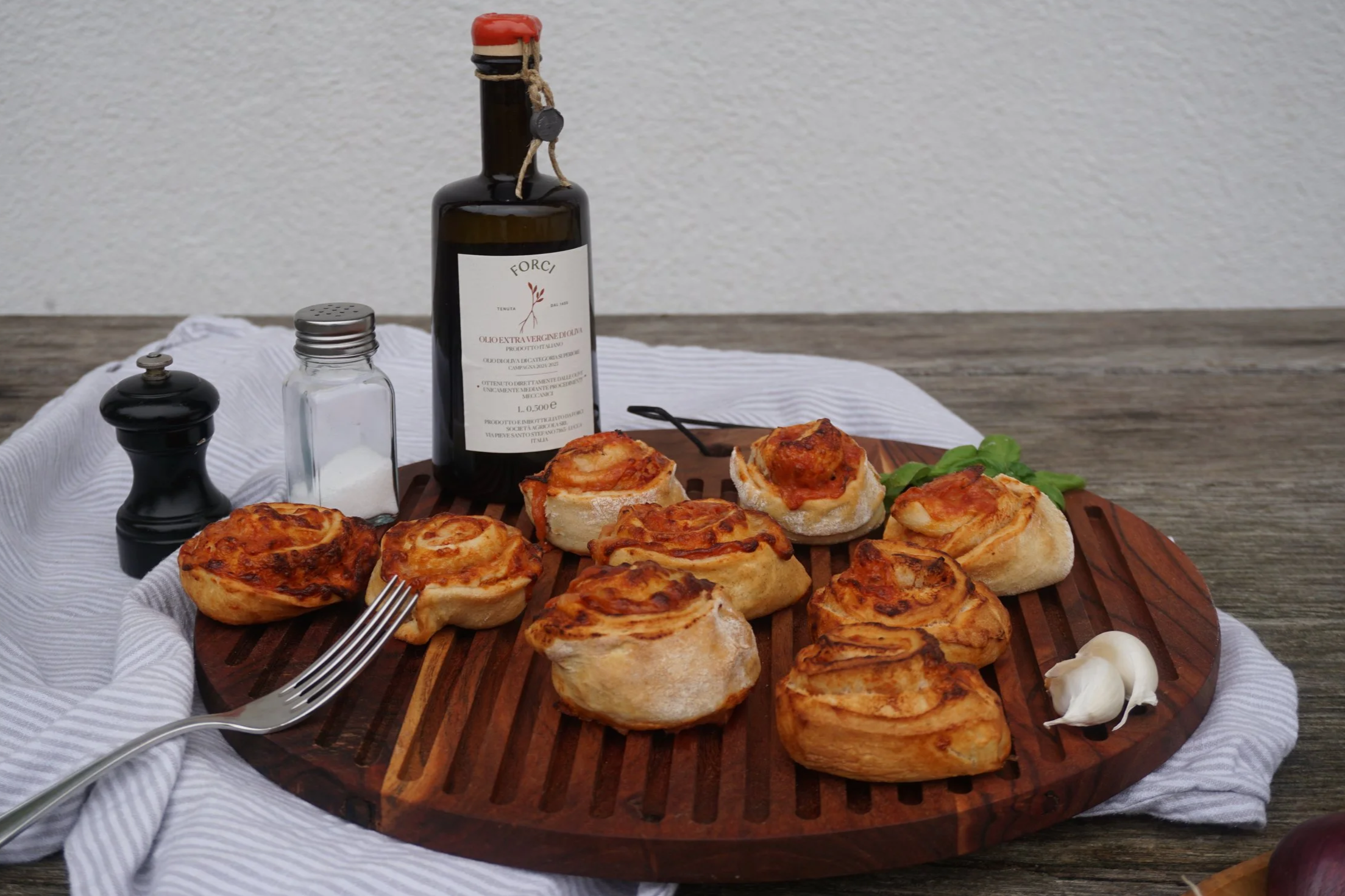 Pizzaschnecken auf einem runden Holzbrett, auf gestreiftem Tuch, mit Salzstreuer, Pfeffermühle und Ölflasche im Hintergrund