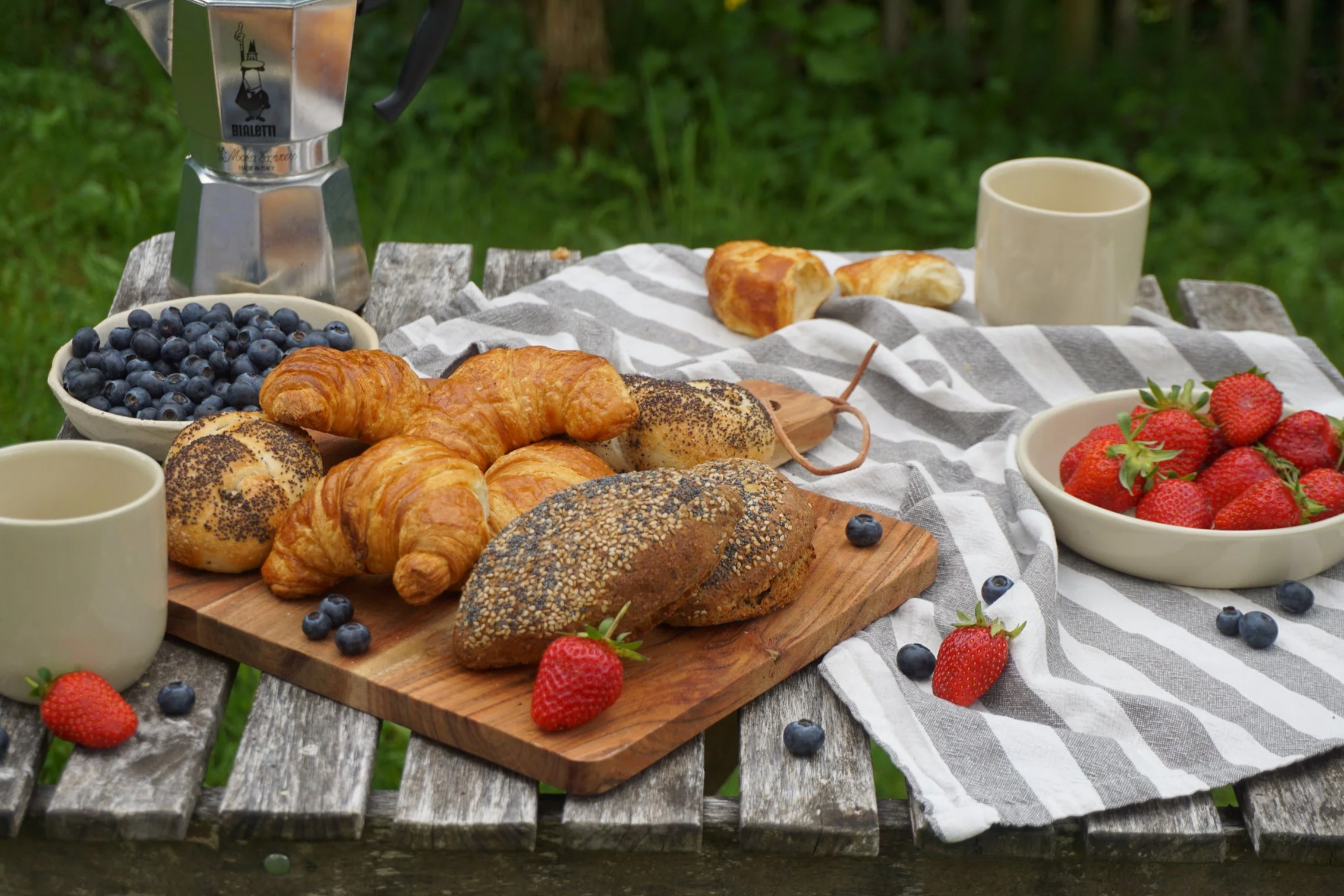 Ein Frühstückstisch im Freien mit Croissants, Brötchen, Beeren, Tassen und einem Espressokocher (Bialetti) auf einem Tisch mit einem weiß, grau gestreiften Tuch.