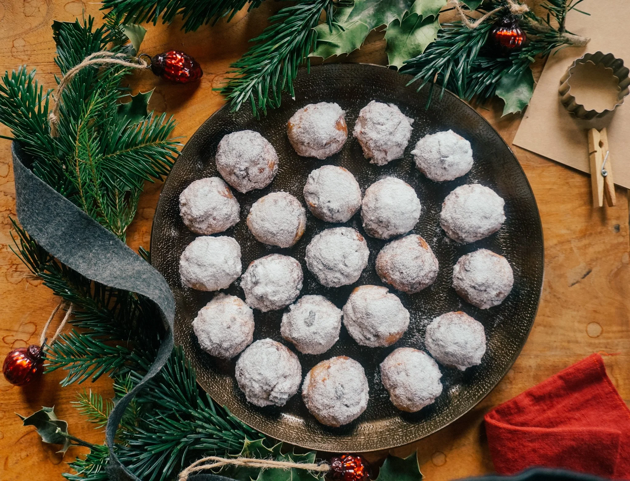 Auf einer schwarzen Platte liegen halbierte Plätzchen, die mit Puderzucker bedeckt sind, umgeben von grünem Weihnachtsdekoration mit Tannenzweigen, Beeren und einem roten Tuch.