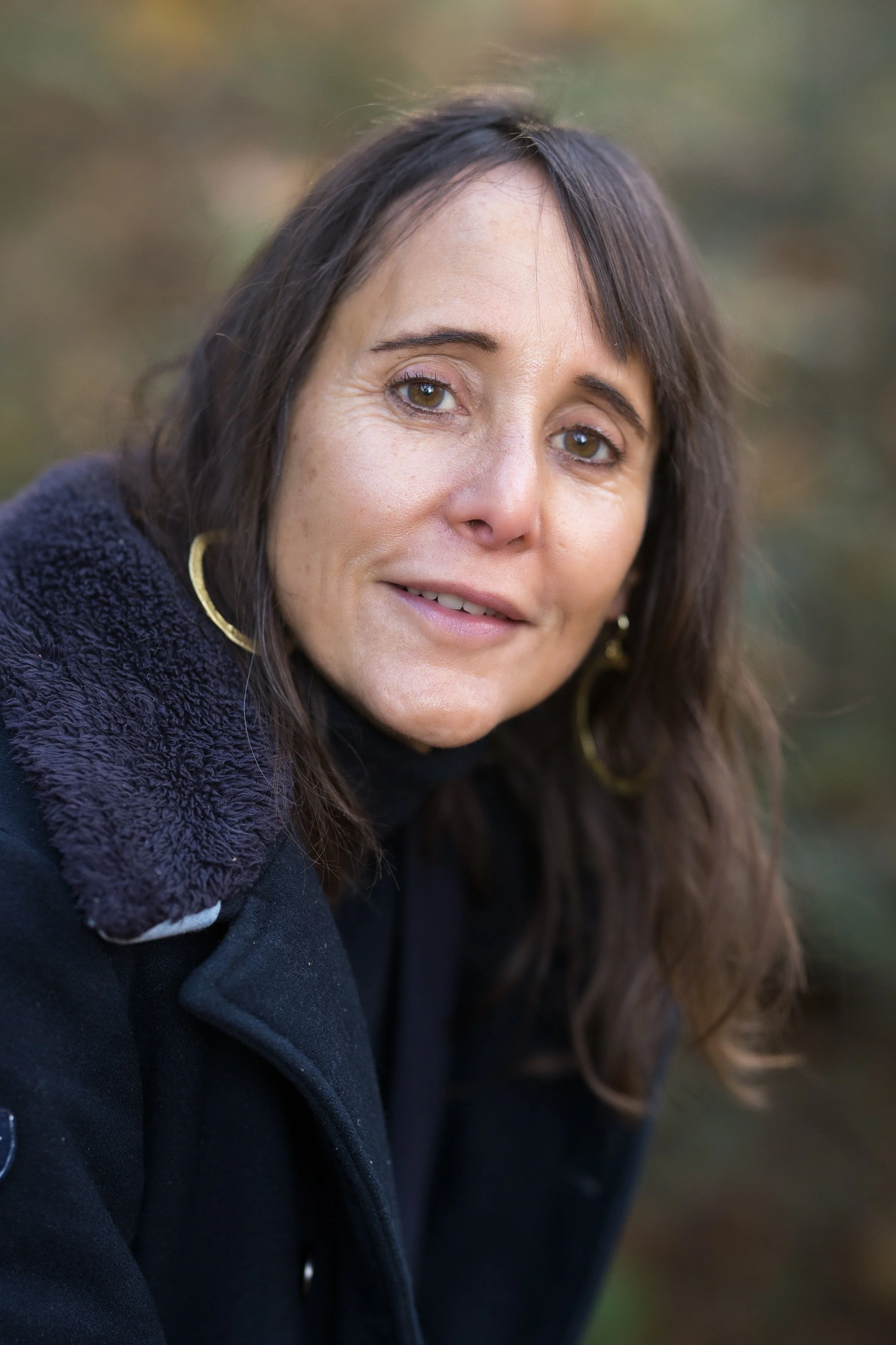 Nahaufnahme einer Frau mit braunen Haaren, Goldohrringen und dunkler Kleidung, vor unscharfem Herbstlaub im Hintergrund.