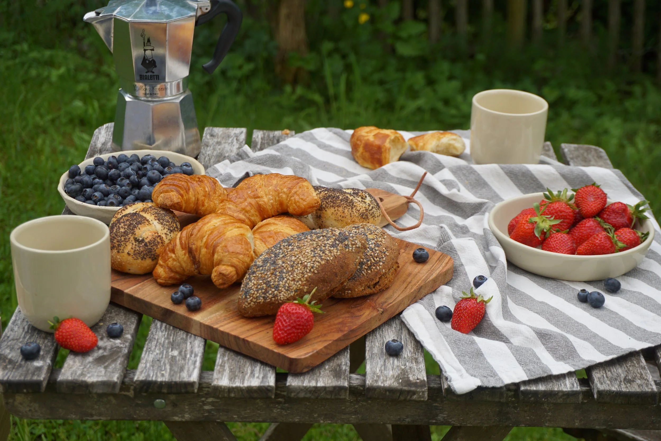 Ein Frühstück im Freien auf einem Holztisch mit einer Zeitung, Joghurt, frischen Erdbeeren, Blaubeeren, Croissants, verschiedenen Brotsorten und einer Espressokanne.