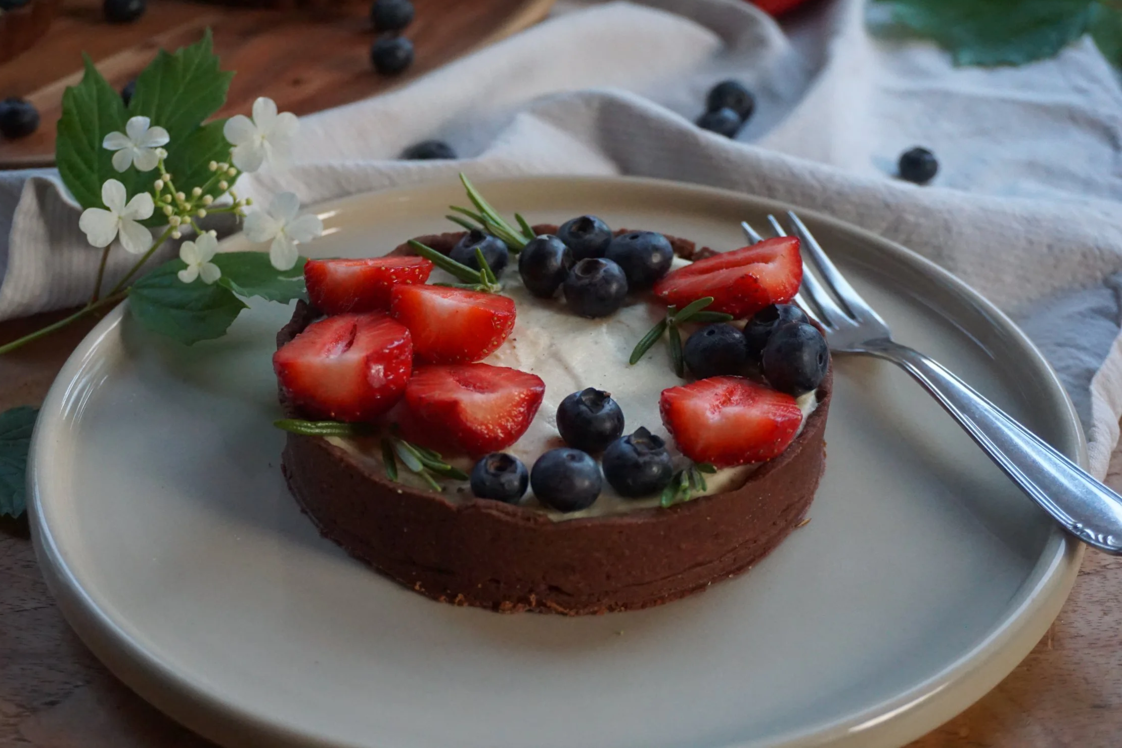 Schokotörtchen, dekoriert mit frischen Erdbeeren, Heidelbeeren, Rosmarin und Holunderblüten auf einem Holztisch.