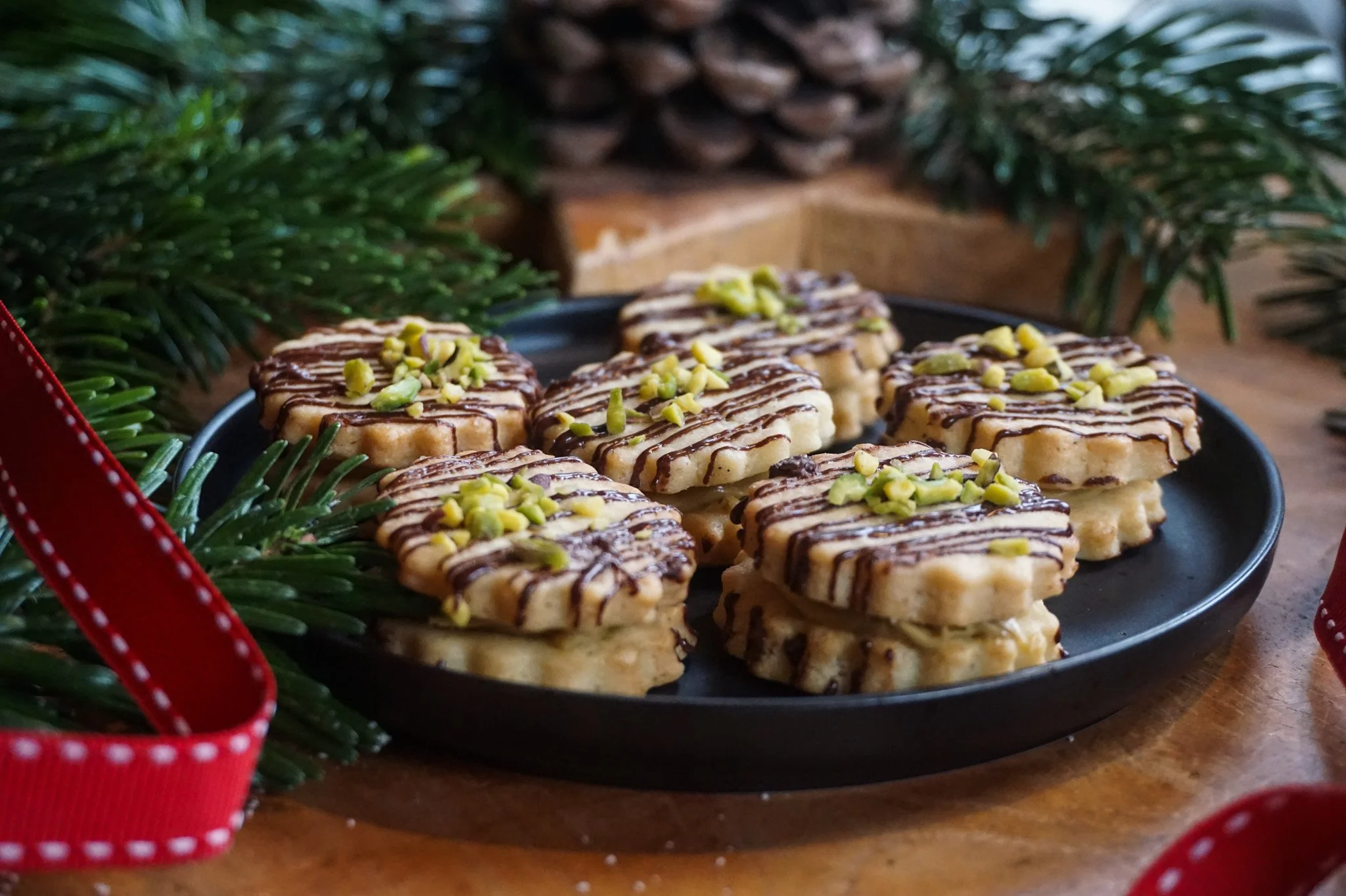 Kekse mit Schokoladensauce und gehackten Pistazien auf einem schwarzen Teller, dekoriert mit Tannenzweigen und roten Geschenkband für Weihnachten.