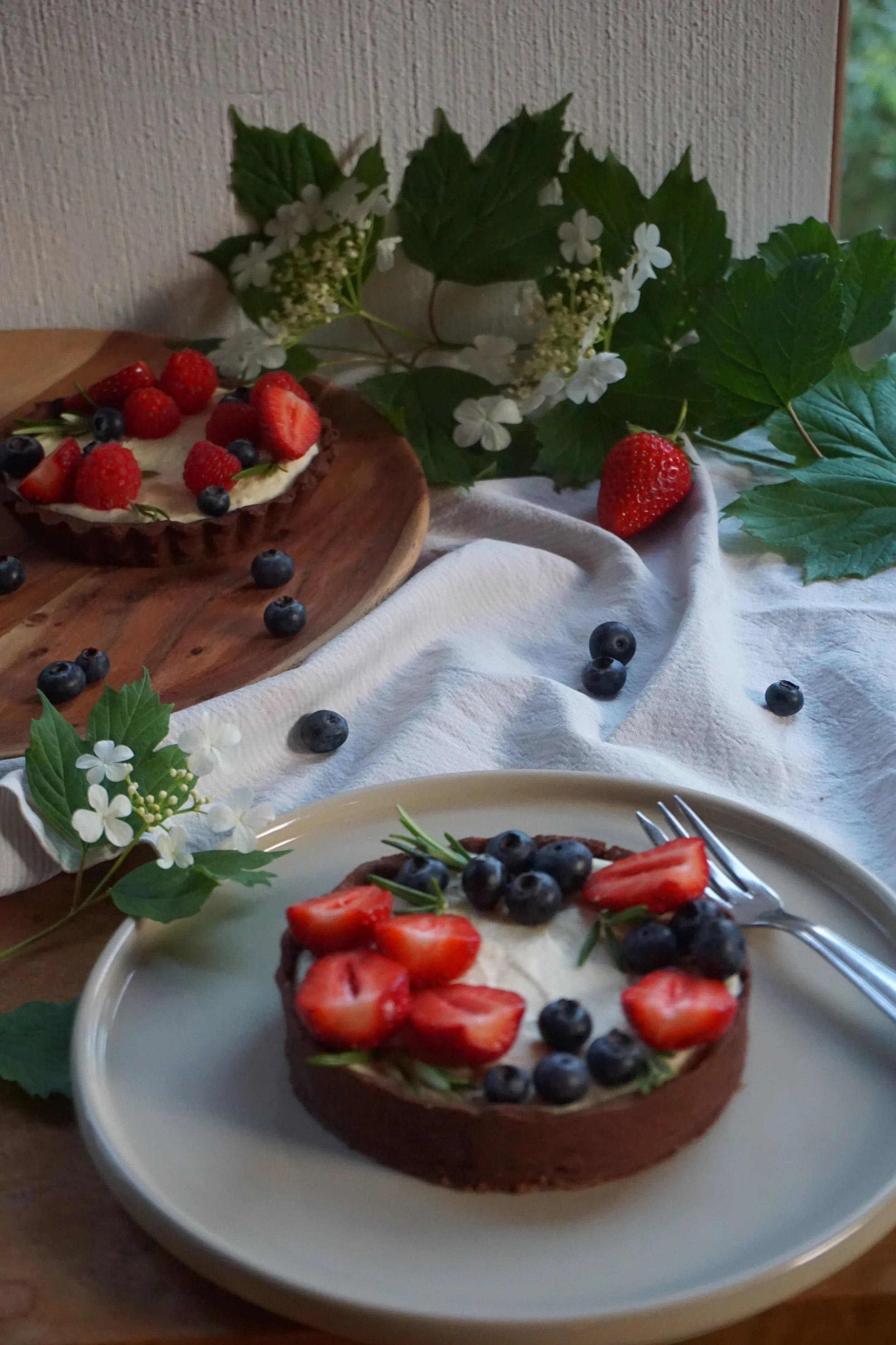 Ein Teller mit Schokoladentörtchen, belegt mit Erdbeeren, Heidelbeeren und Rosmarin, auf einem weißen Teller. Hintergrund mit grünen Blättern und weißen Blumen.