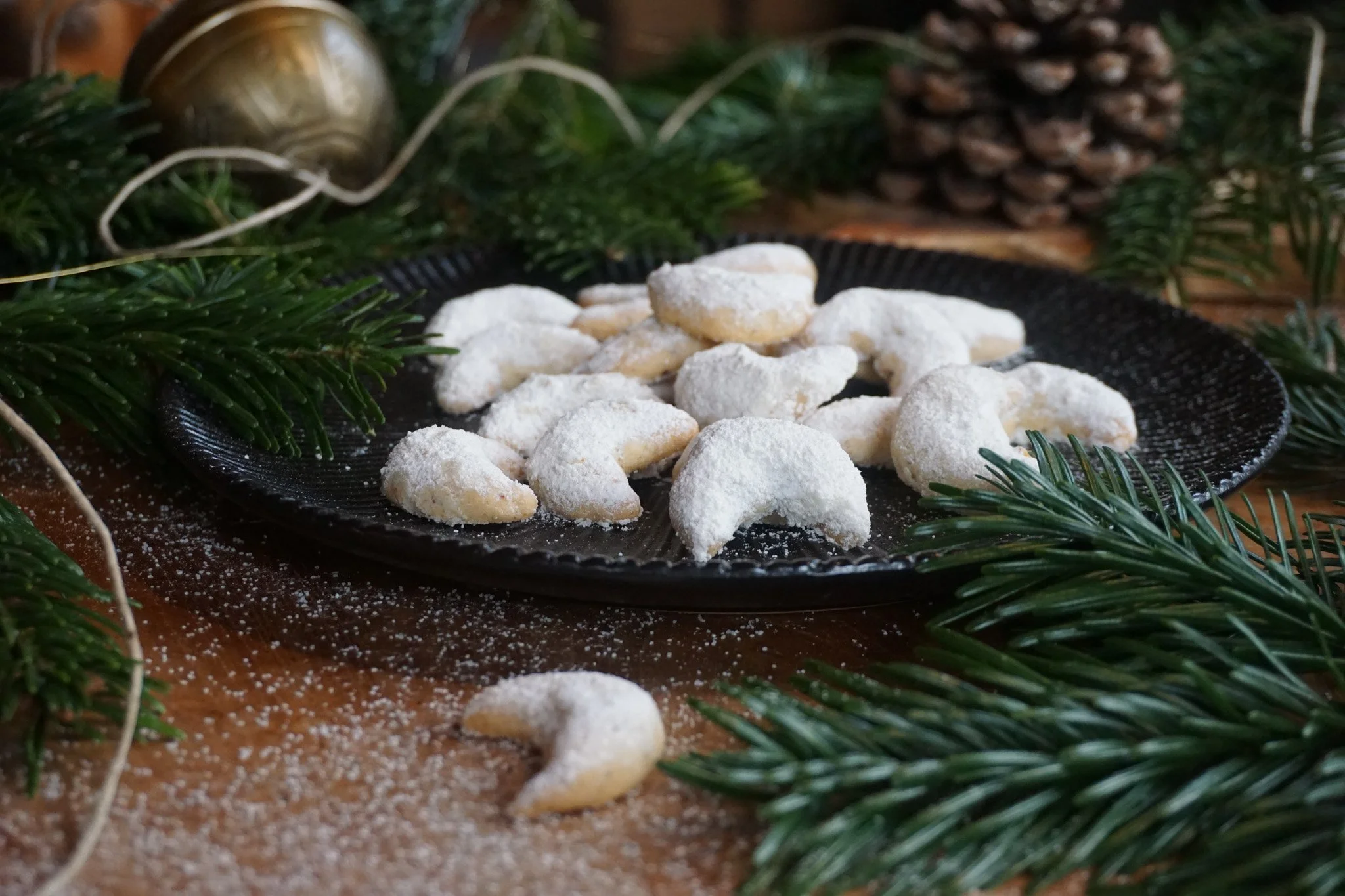 Weiße Kekse in Form von Schneefamilien liegen auf einem schwarzen Teller, umgeben von Tannenzweigen und einer goldenen Weihnachtskugel, mit etwas Puderzucker bestäubt.
