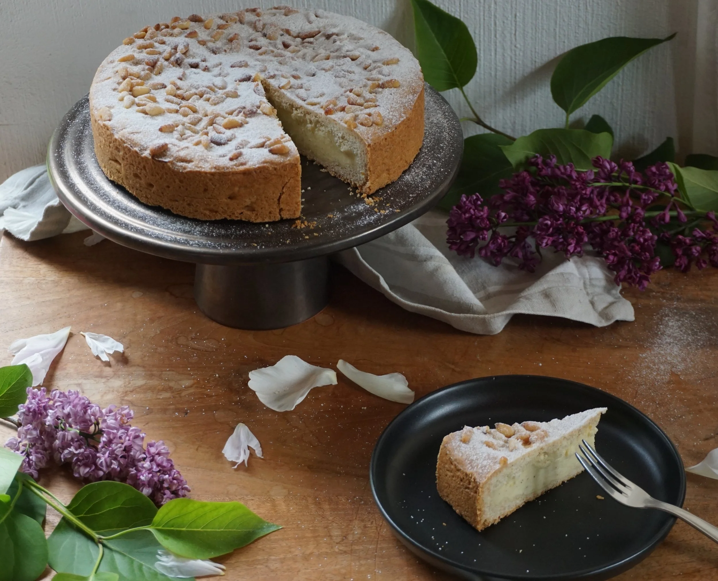 Italienischer Kuchen mit Puddingfüllung und Pinienkernen und Puderzucker bedeckt. Mit einzelnem Kuchenstück im Vordergrund auf einem schwarzen Teller angerichtet. Mit Flieder dekoriert