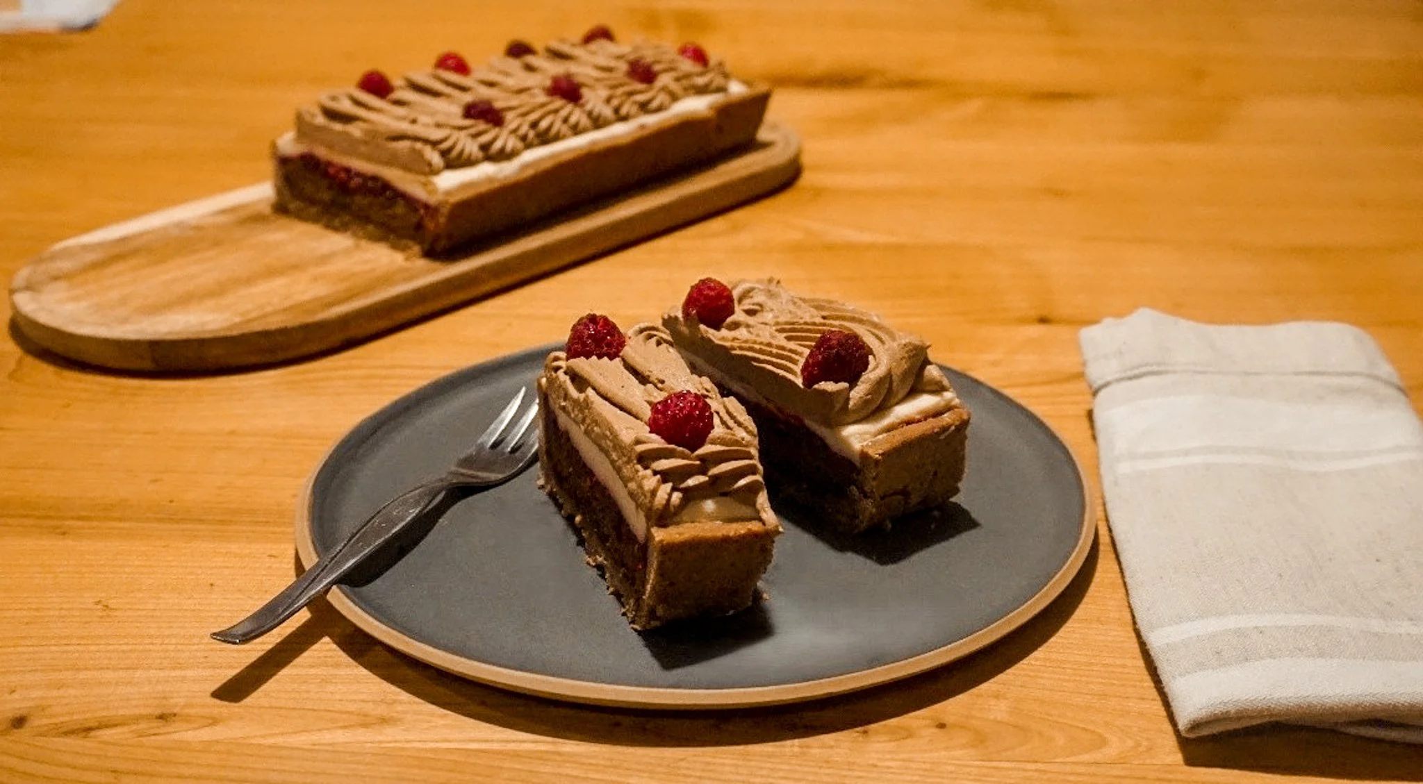 Zwei Stücke Dessert mit roter Beere auf einem schwarzen Teller, davor ein Messer und Gabel, im Hintergrund ein Tortenstück auf einem Holzbrett, rechts eine weiße Stoffserviette auf einem Holztisch.