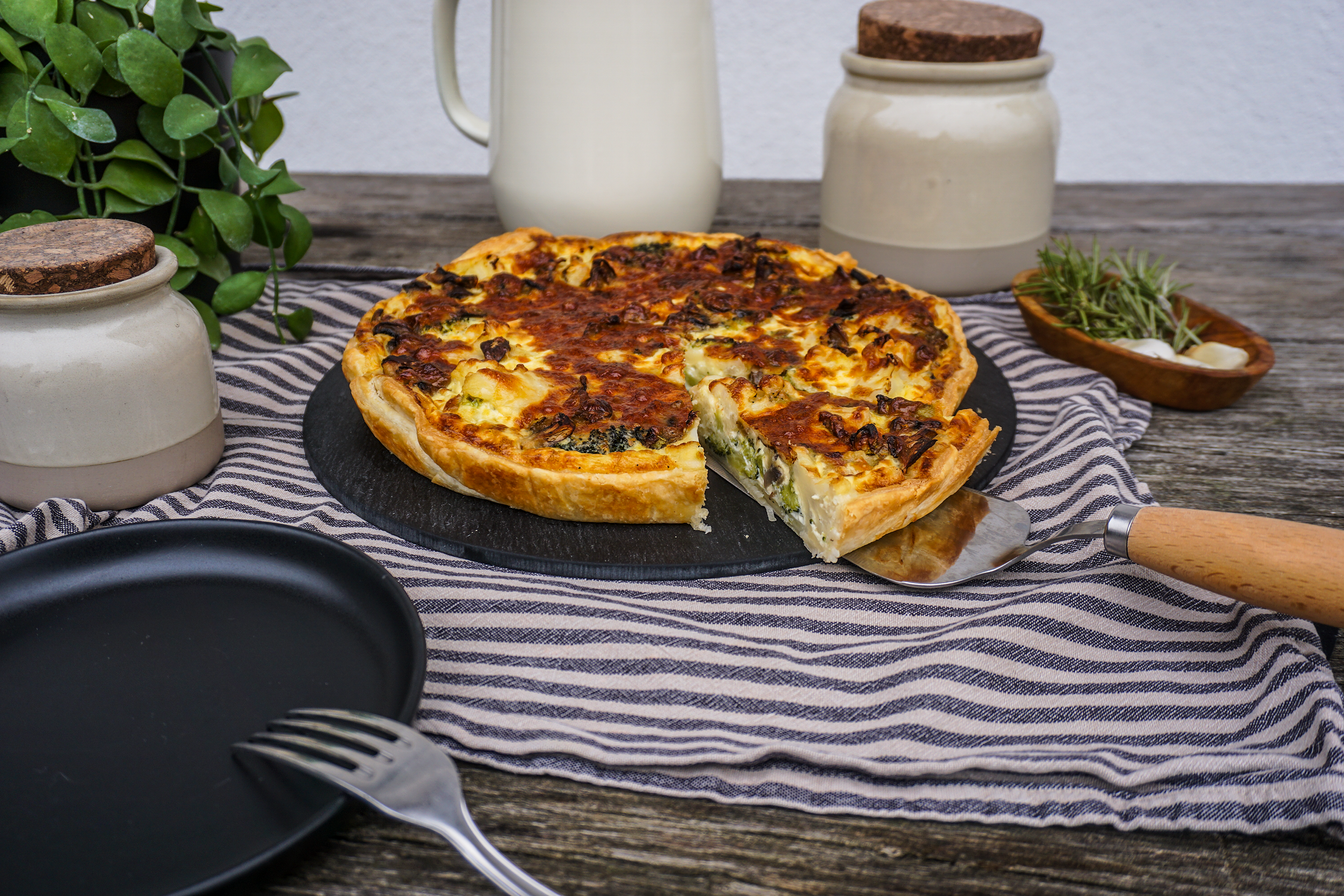 Italienische Pizza mit Käse und Gemüse auf schwarzener Platte, serviert auf grobem Holztisch mit Besteck, Geschirr und Dekoration.