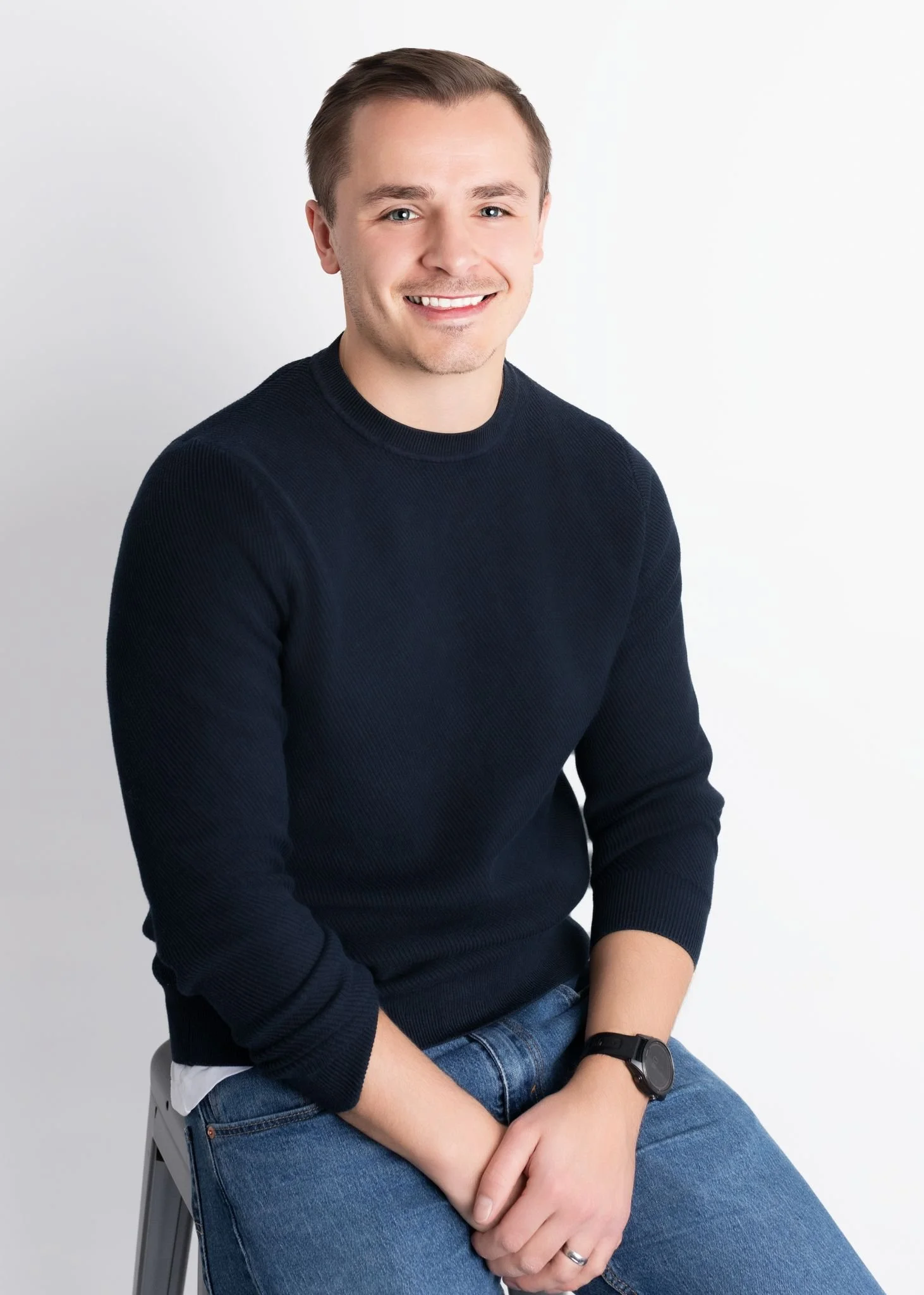 A young man with short brown hair, wearing a black sweater, blue jeans, and a black watch, sitting against a plain white background and smiling at the camera.