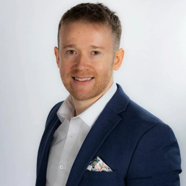 A man with short light brown hair and a beard, wearing a dark blue suit jacket, white shirt, and patterned pocket square, smiling against a plain white background.