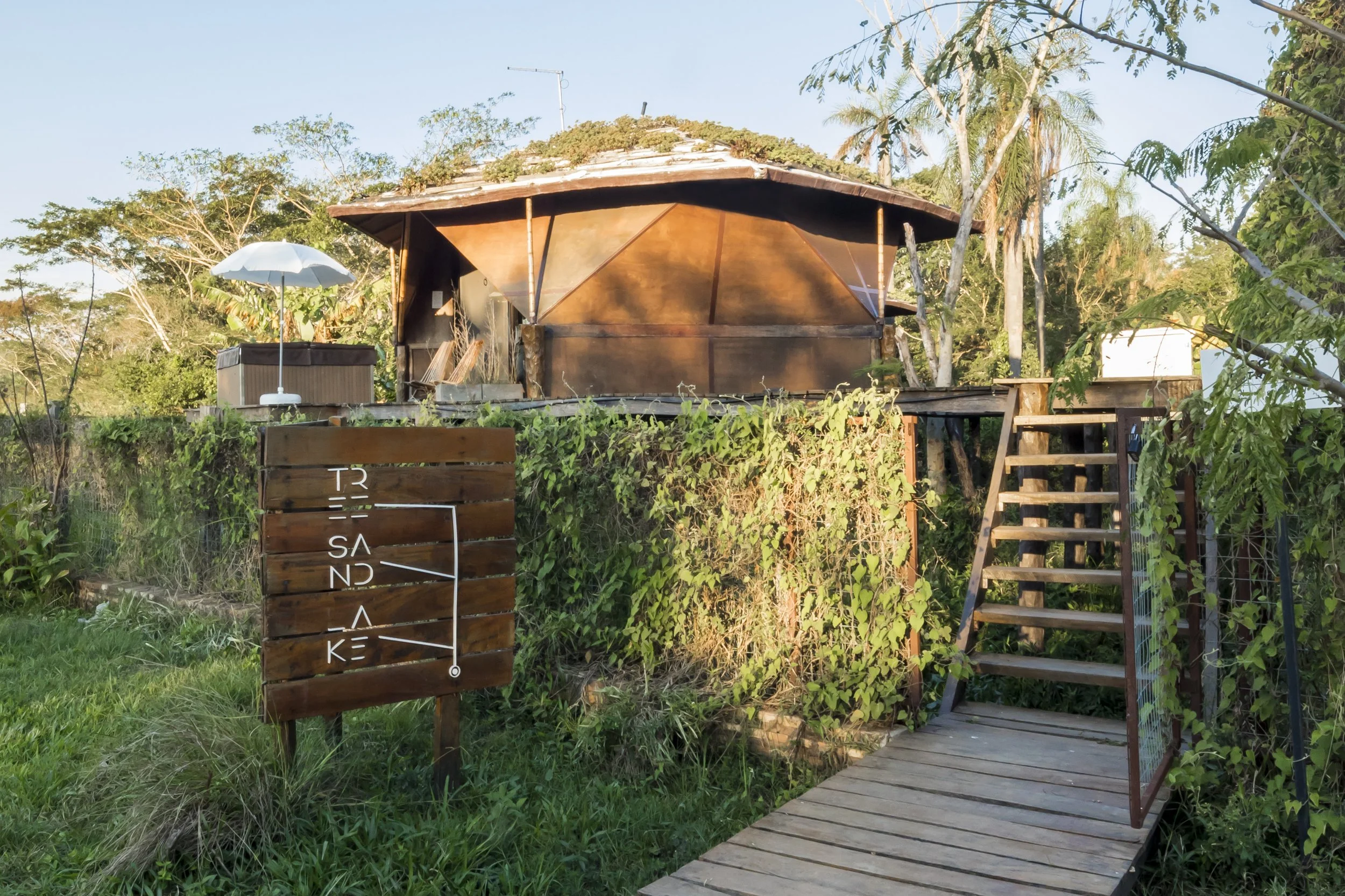 PRO ARQUITECTURA PARAGUAY architecture design PROSA
Casa ecológica de madera en un entorno natural rodeada de vegetación, con un cartel de madera y un camino de madera con una pequeña escalera de acceso.
