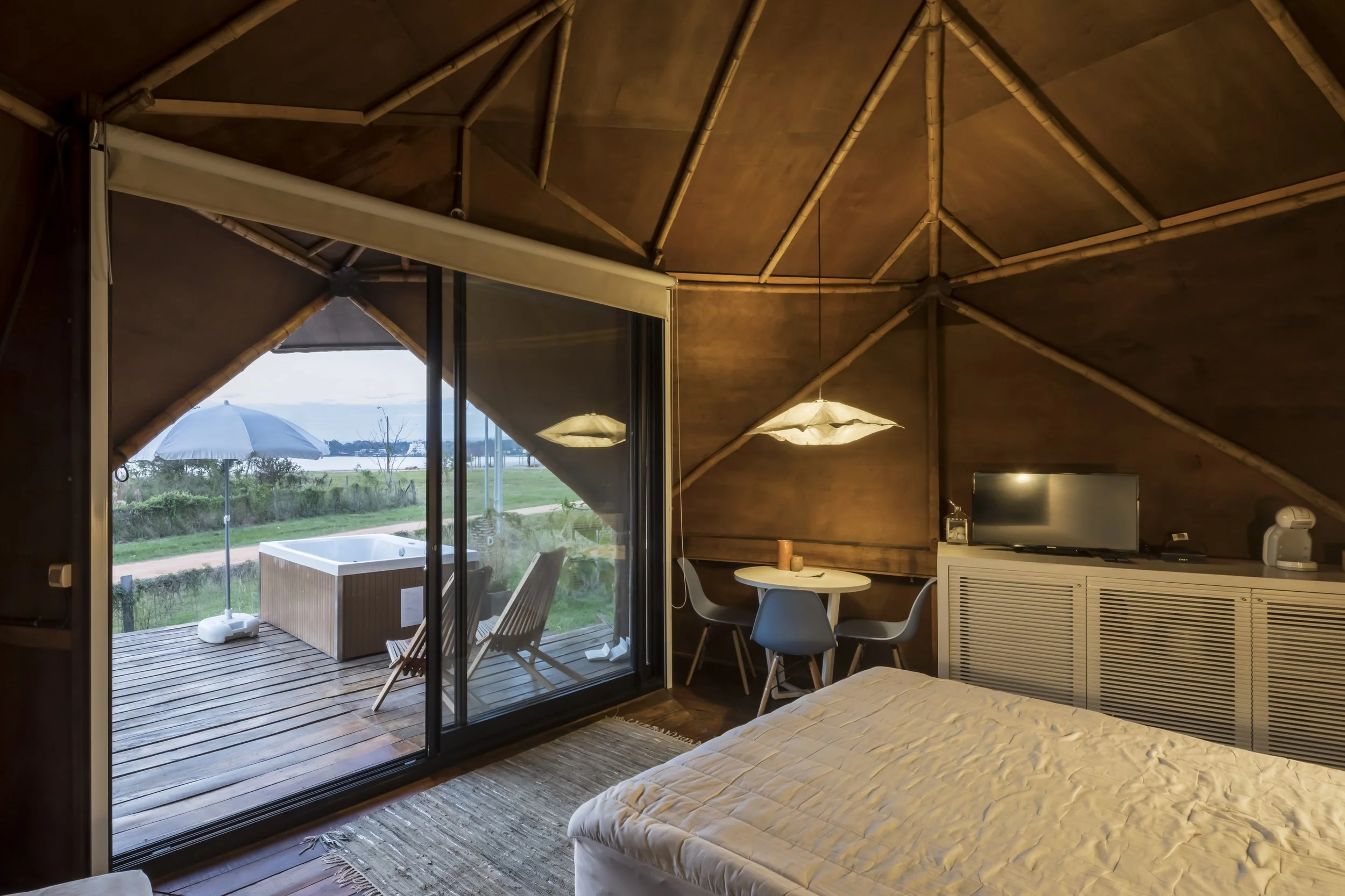 PRO ARQUITECTURA PARAGUAY architecture design PROSA
Interior de una habitación de hotel con techo de madera, cama, televisor, mesa con sillas, lámpara y vista a un balcón con jacuzzi, paraguas y paisaje natural.