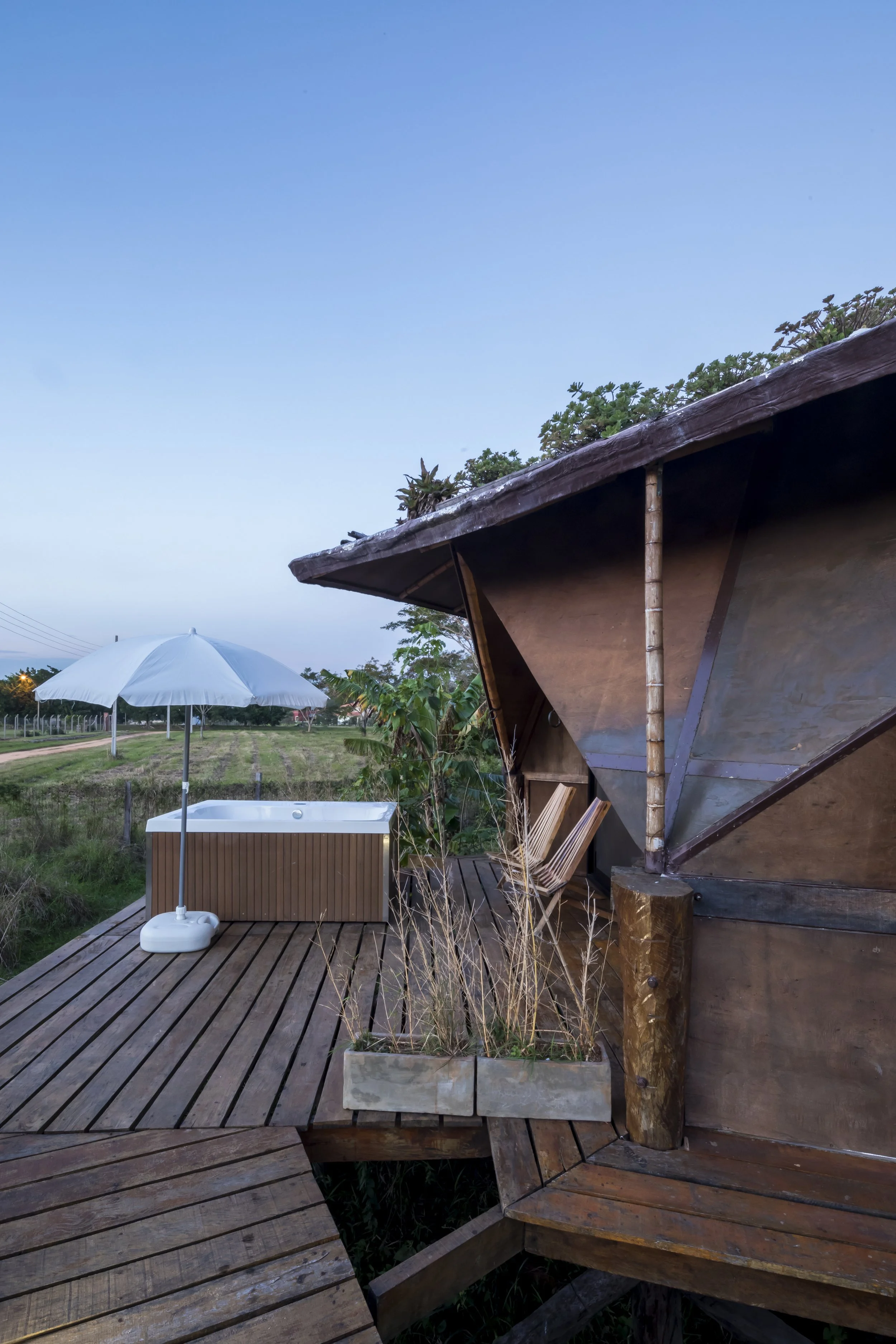 PRO ARQUITECTURA PARAGUAY architecture design PROSA
Una terraza de madera con una bañera, una sombrilla, sillas y plantas, rodeada de naturaleza en un entorno rural durante el atardecer.